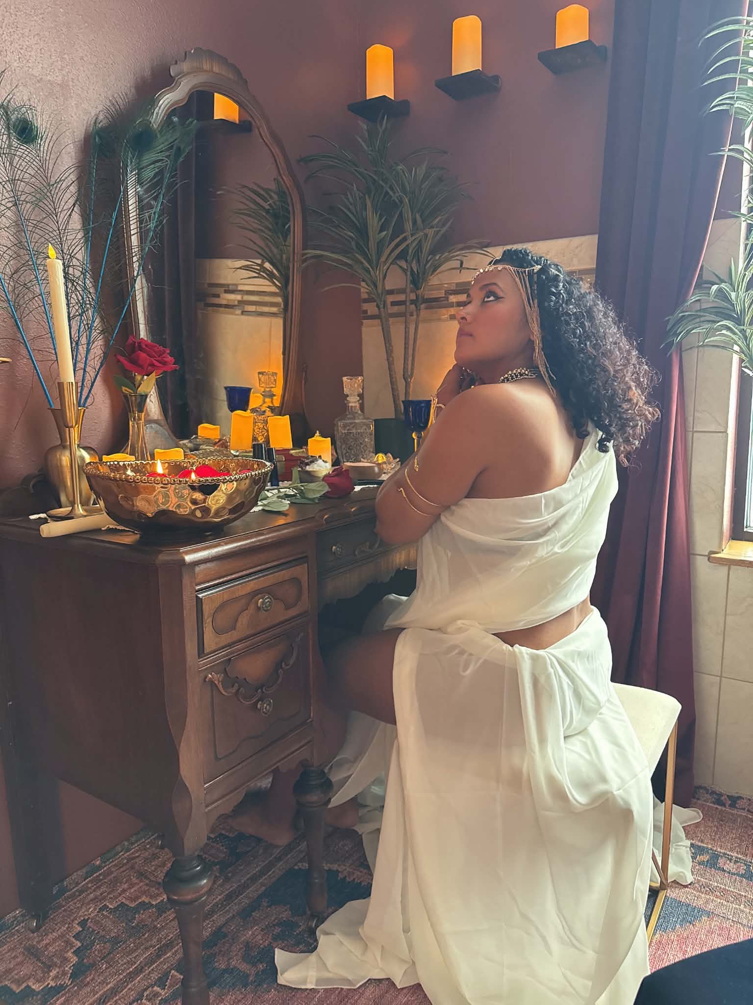 Woman with curly hair in white dress sitting at a decorated table with candles and flowers, in a warmly lit room.