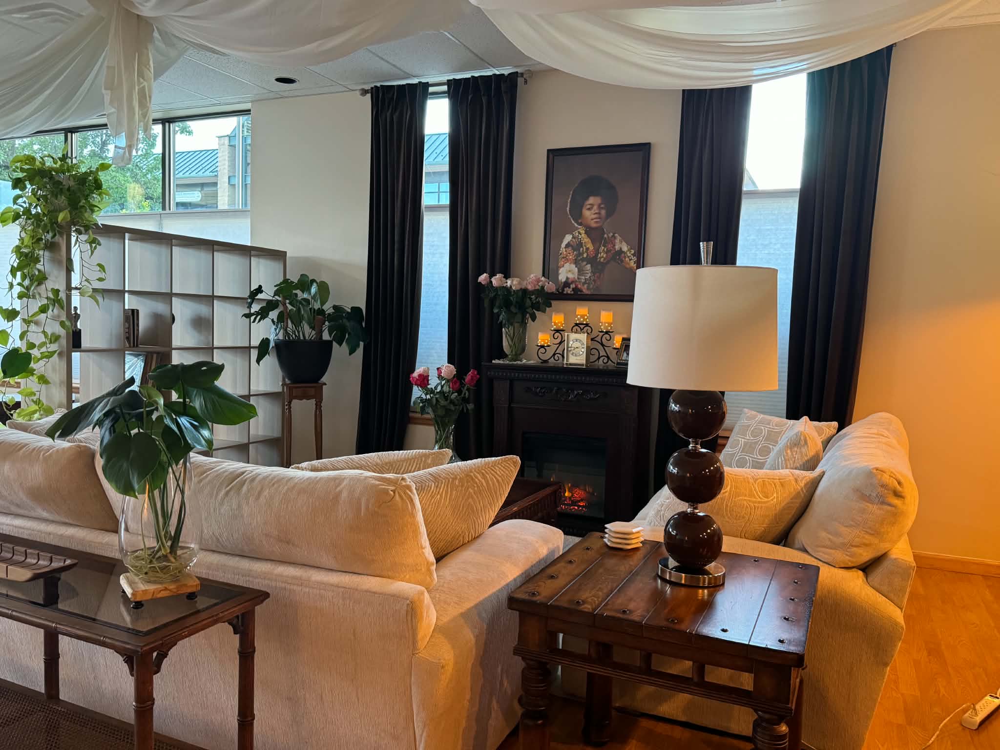 Living room with beige sofas, wooden tables, a lamp, framed artwork, and large windows with dark curtains.
