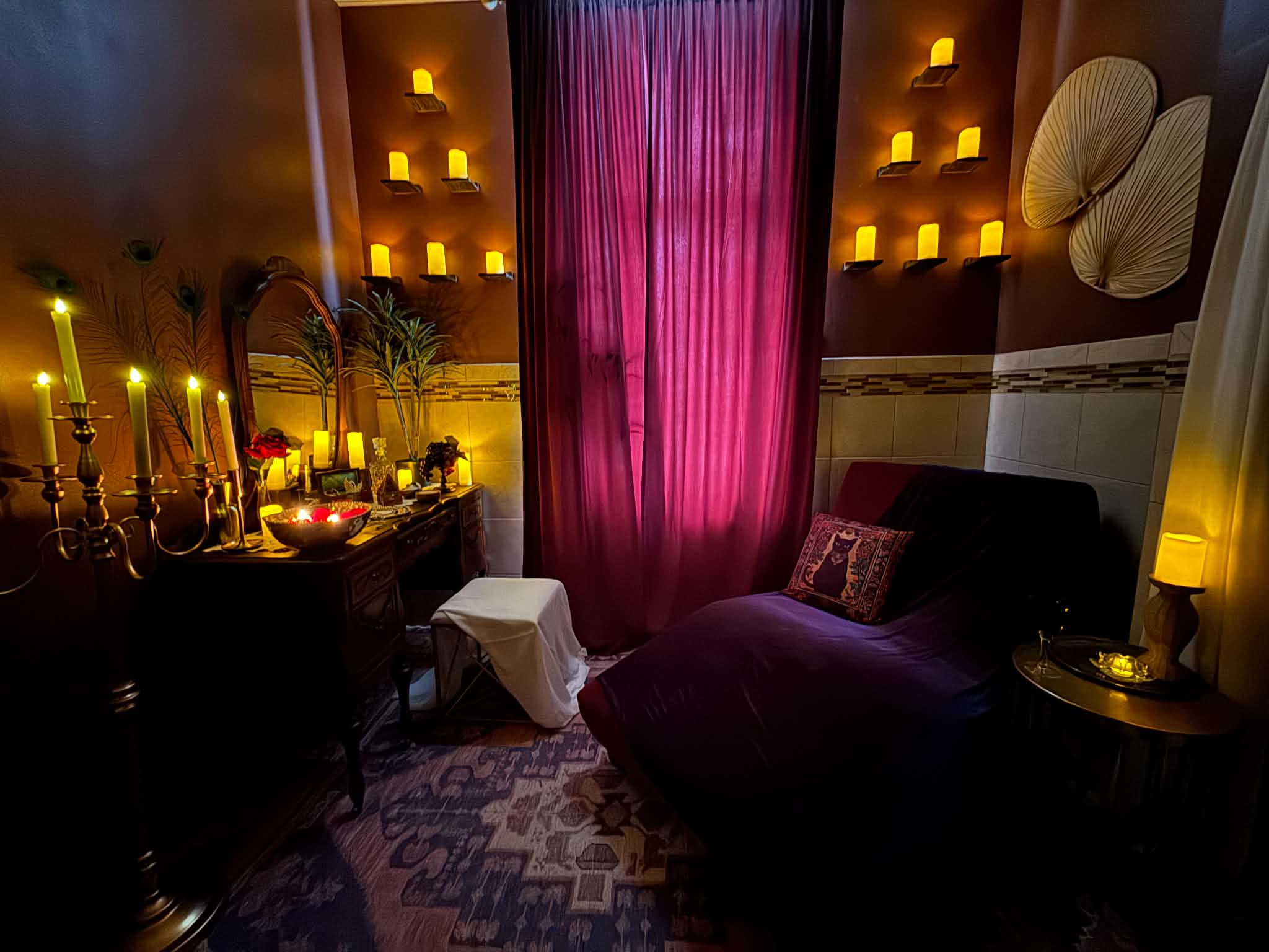Cozy room with purple curtains, a dark sofa with a pillow, and warm lighting from candles and lamps.