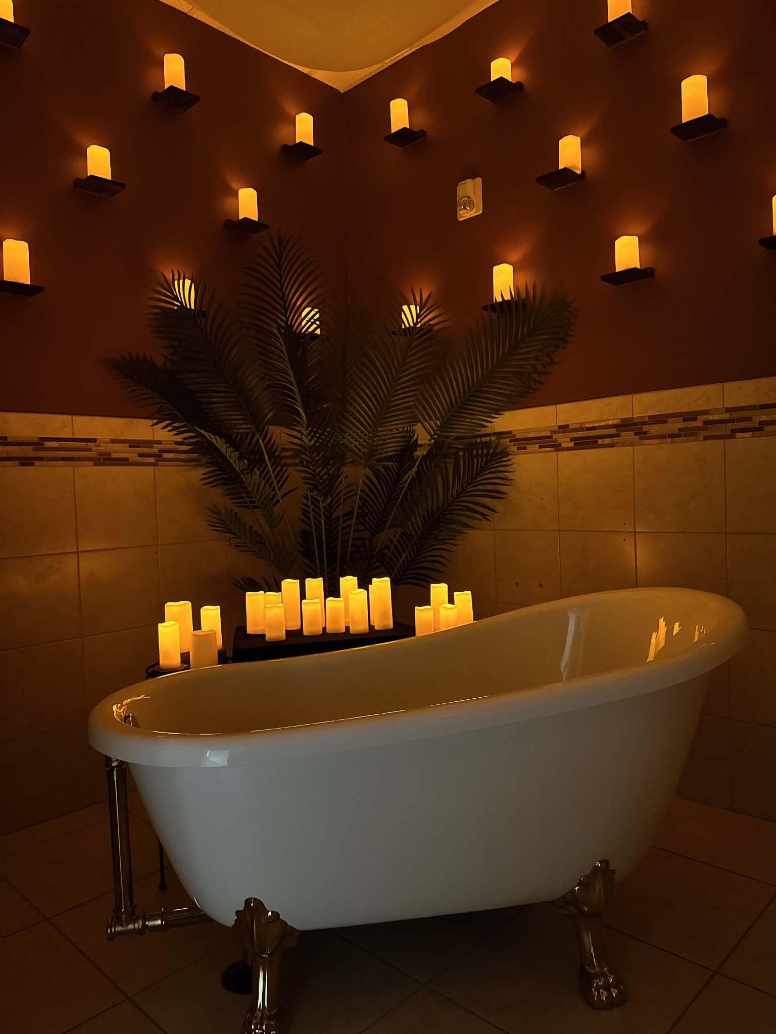 Bathtub in a dimly lit room with numerous candles and a large plant behind it.