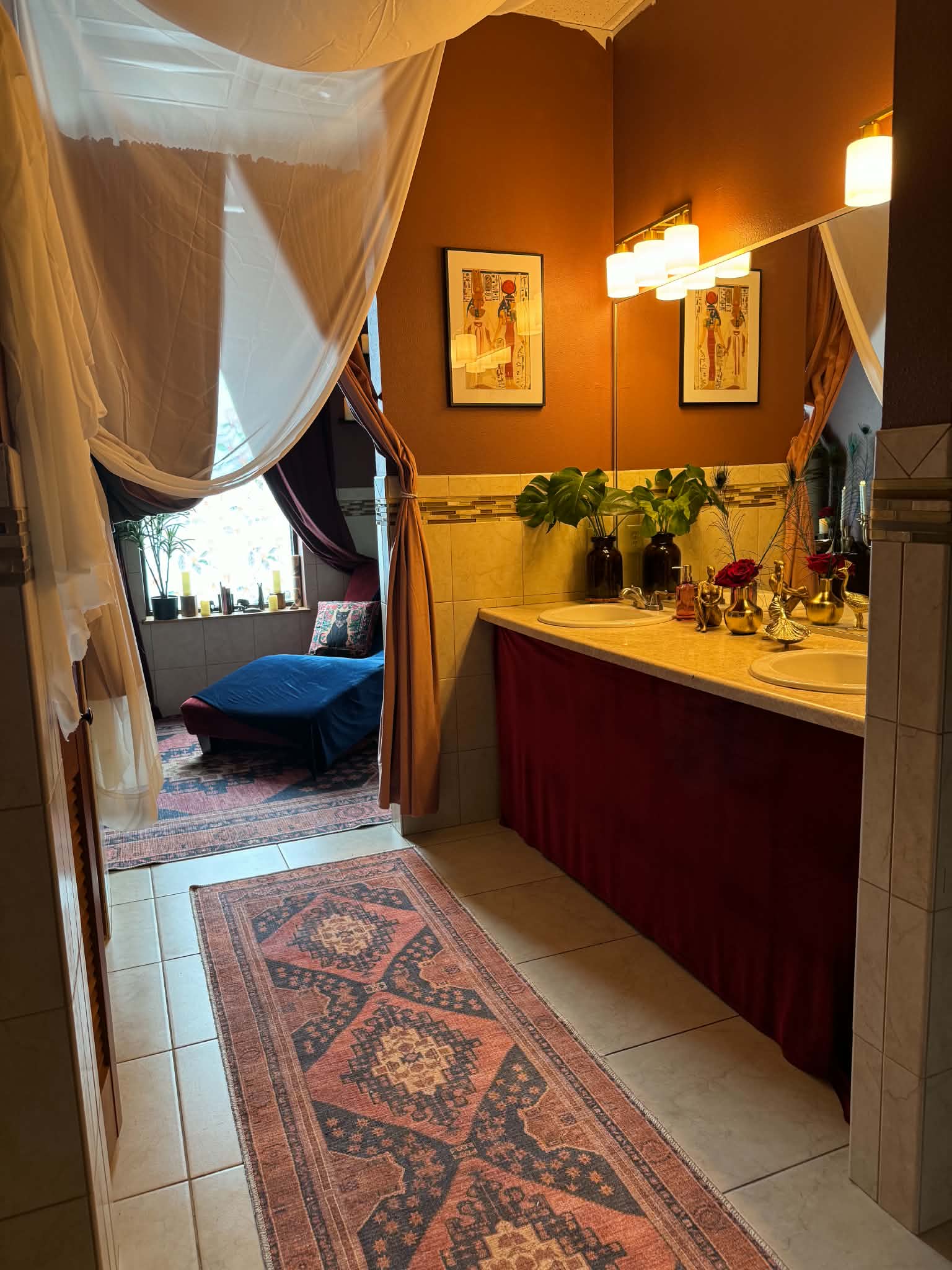 Bathroom with a sink, mirror, and wall-mounted lights, decorated with plants and framed artwork, with a doorway leading to a living area.