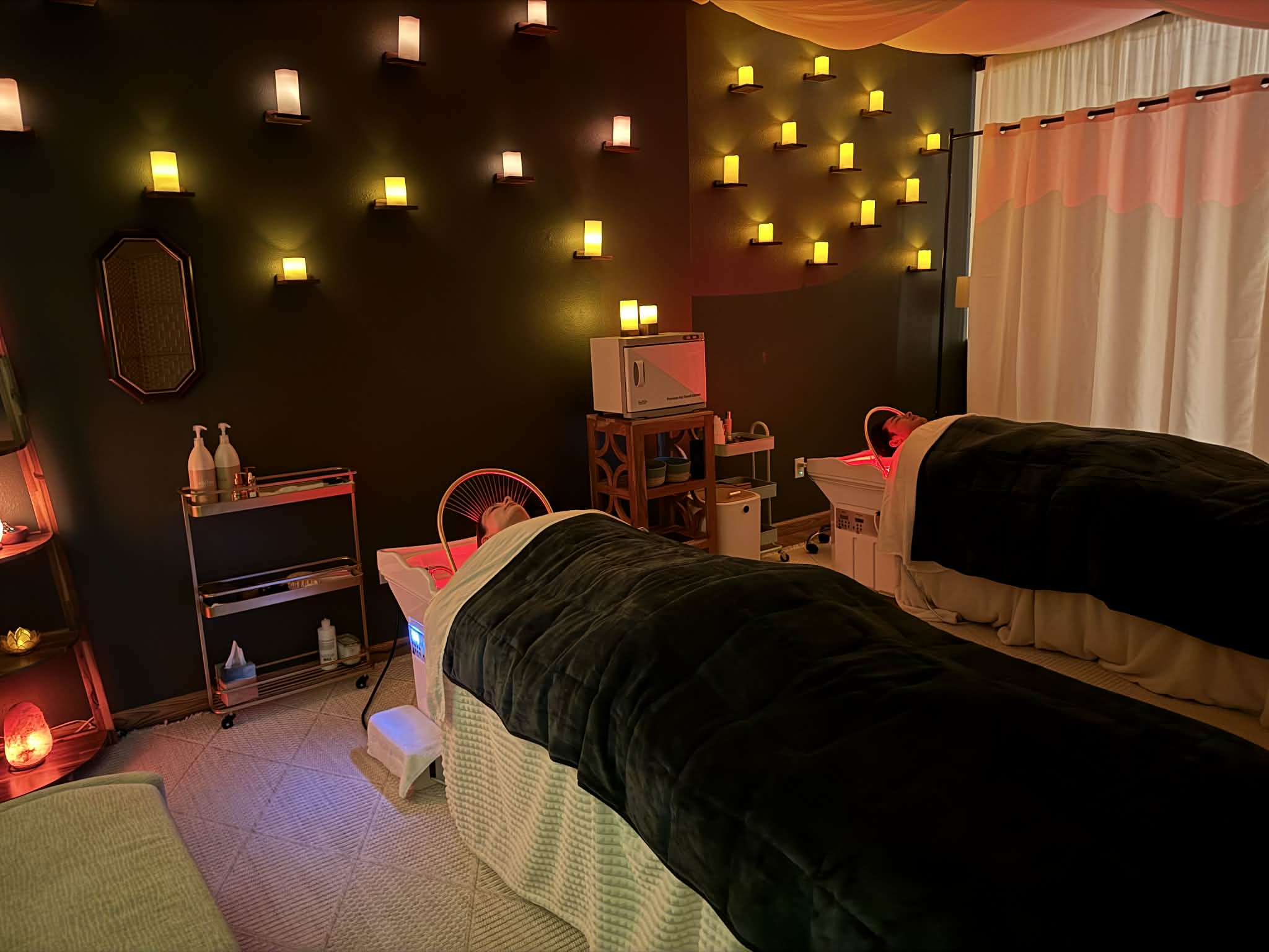 Spa room with two massage beds covered with black sheets, warm lighting, and a dark wall with small illuminated squares.
