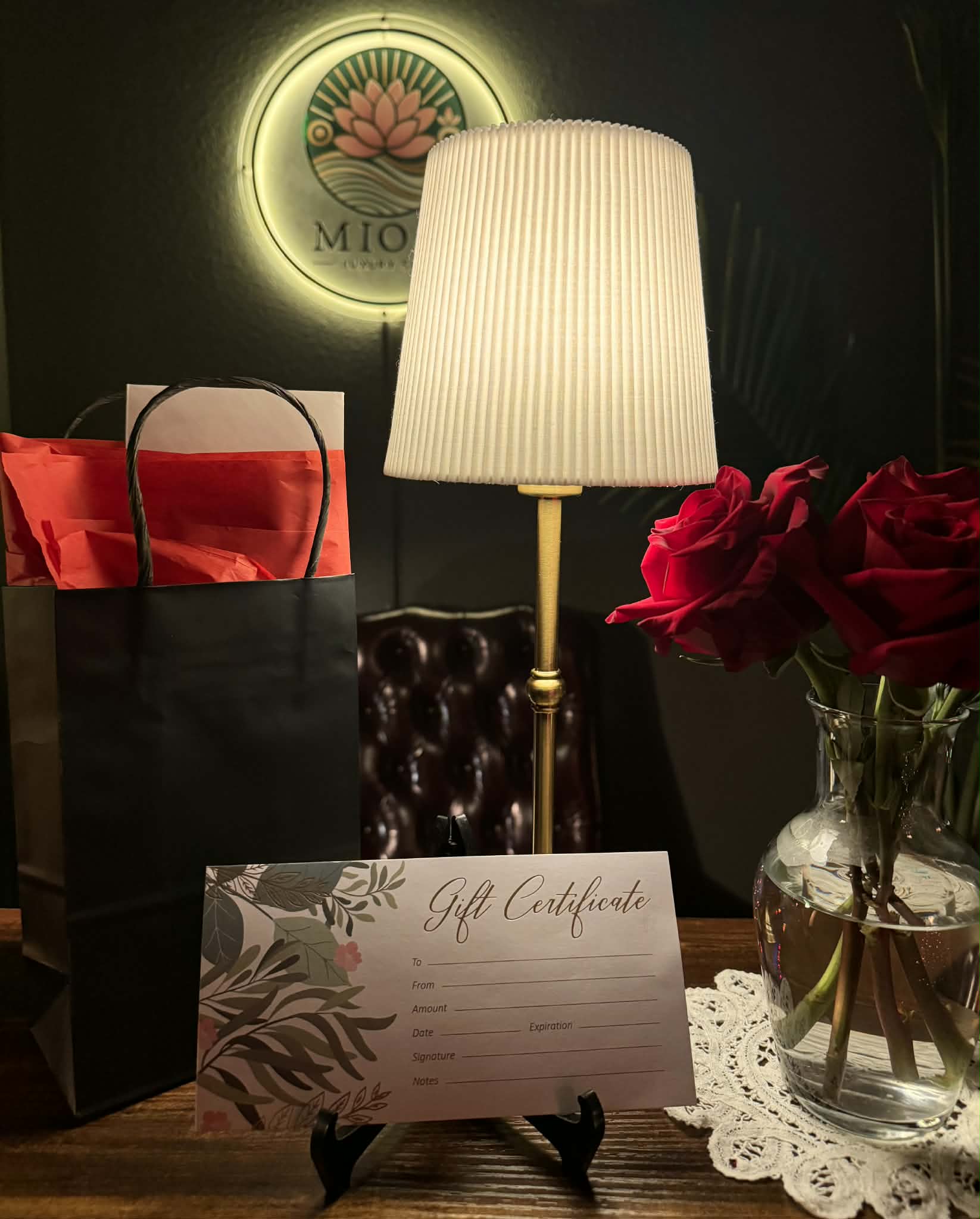 Table with a gift certificate, a lamp, a vase of red roses, and a gift bag, in a dimly lit setting.