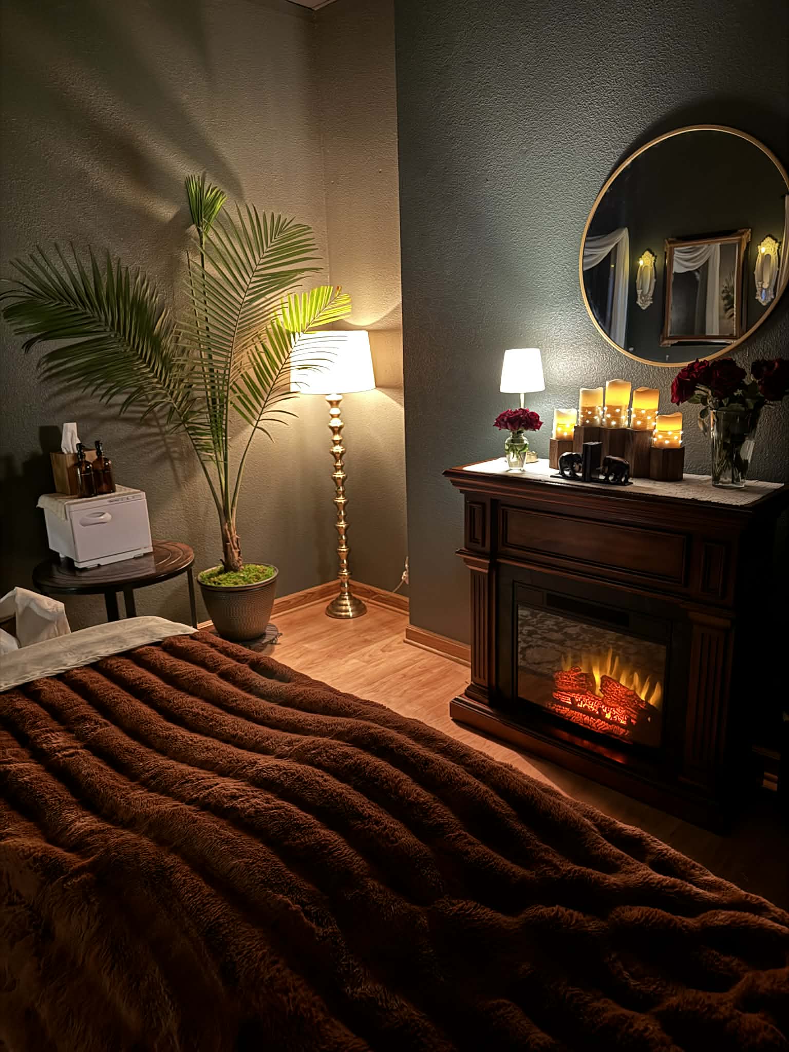 Cozy bedroom corner with a bed, potted plant, standing lamp, and a fireplace with a mirror above, warm lighting.