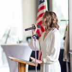 Woman speaking into microphone at podium with American flag in background, wearing white dress, smiling, in a bright room.