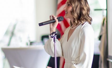 Woman speaking into microphone at podium with American flag in background, wearing white dress, smiling, in a bright room.