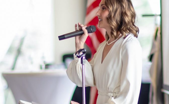 Woman speaking into microphone at podium with American flag in background, wearing white dress, smiling, in a bright room.
