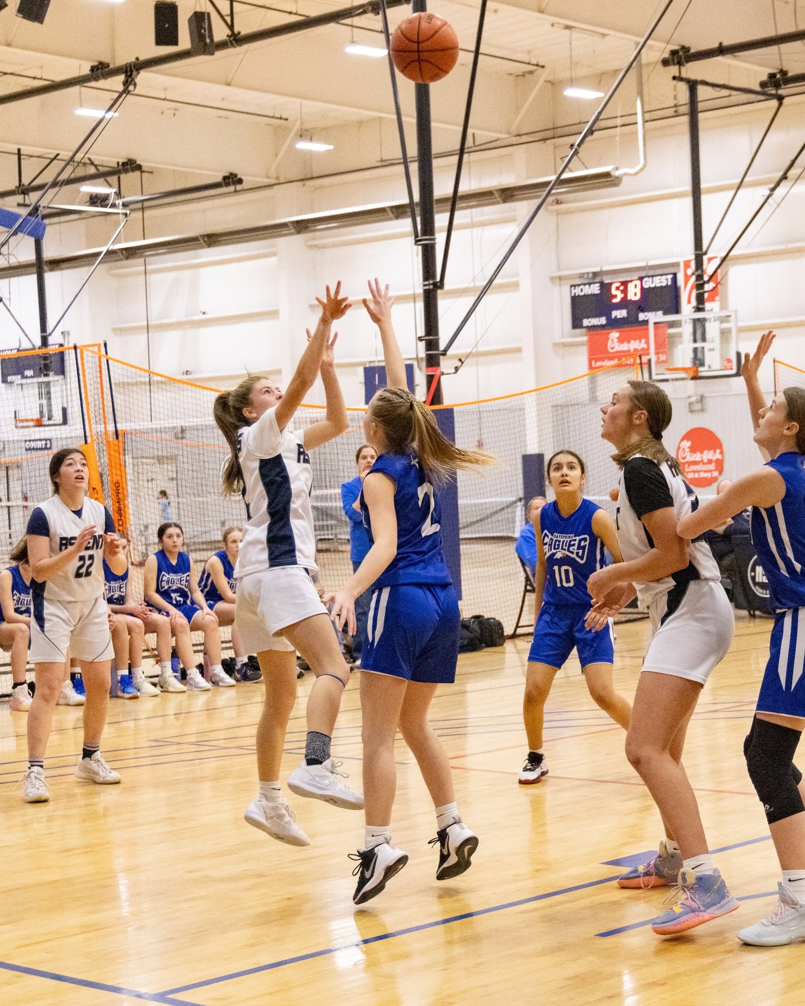 Girls playing basketball on an indoor court, some jumping to shoot or block, others watching or preparing to play.