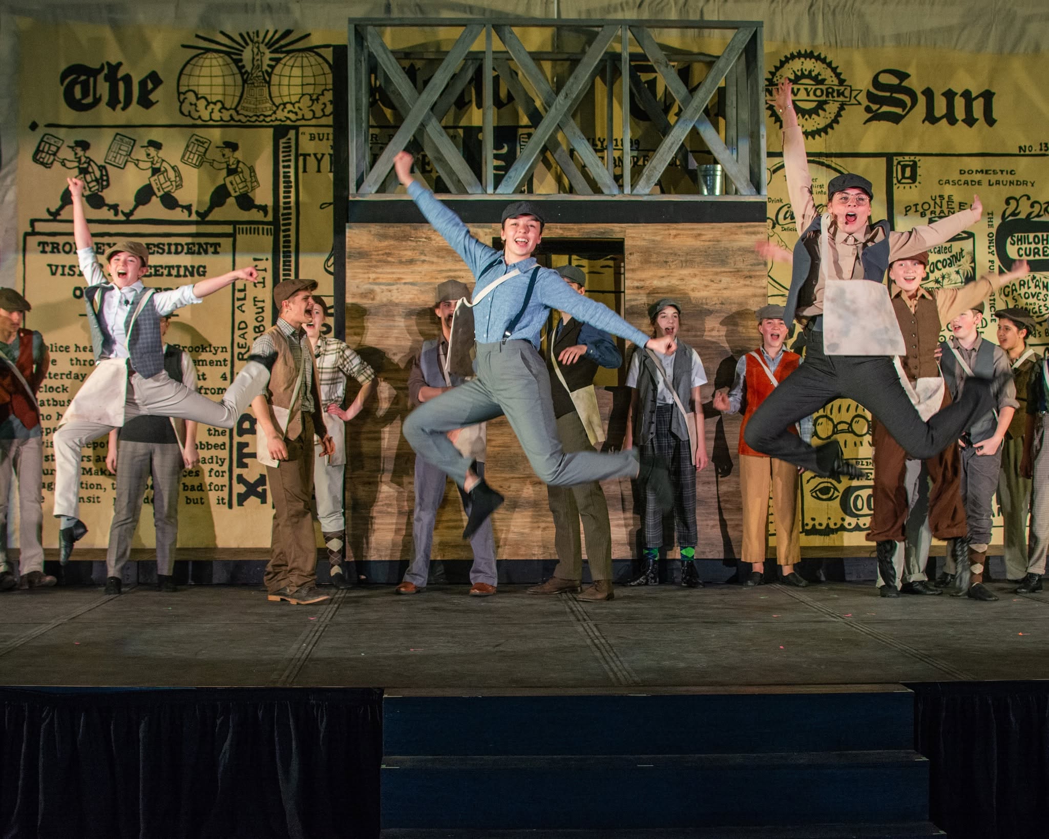 Theater performance with dancers in vintage costumes on stage, background resembling a newspaper, wooden structure in center, lively scene.