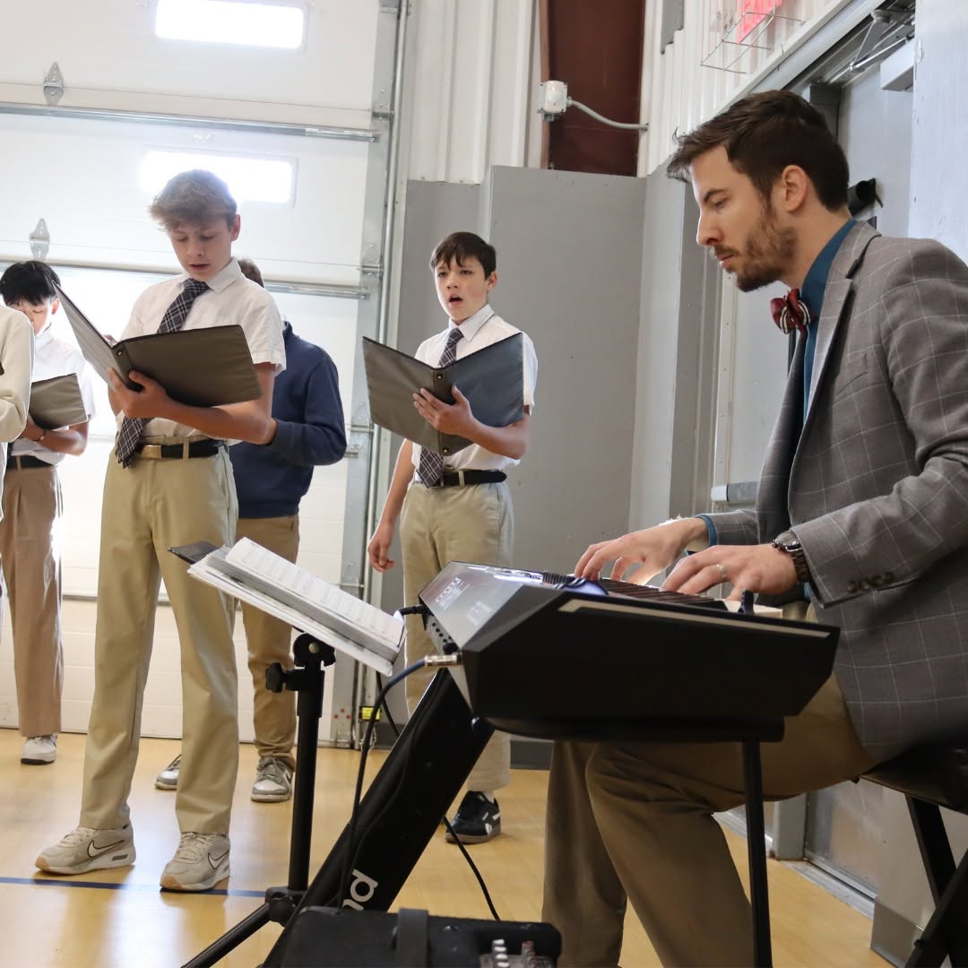 A man playing keyboard with four boys standing nearby, holding books, in a room with large windows and a garage door.