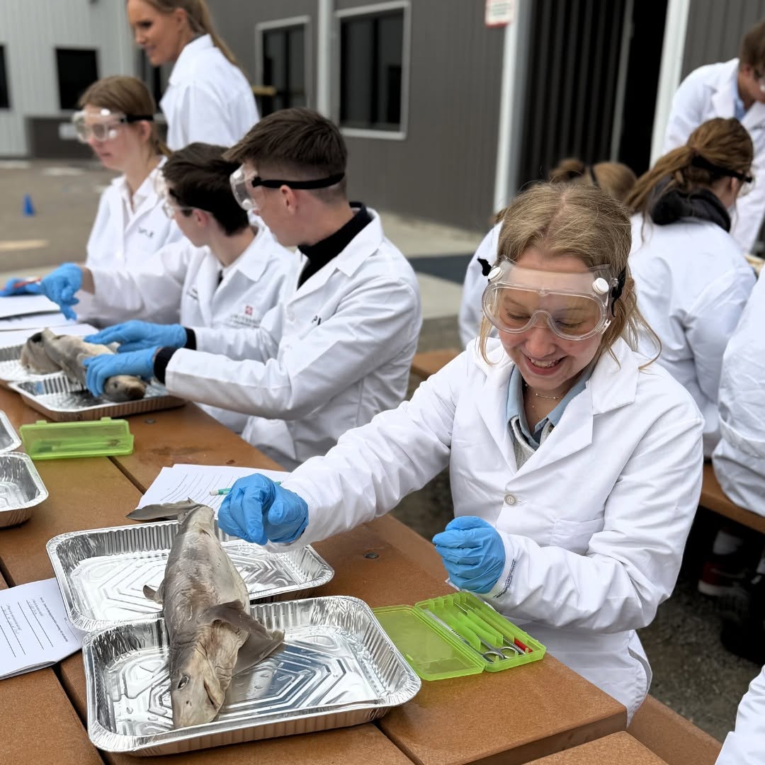 Group of students in lab coats and goggles working with a fish on a table outdoors, smiling and handling the fish with gloves.