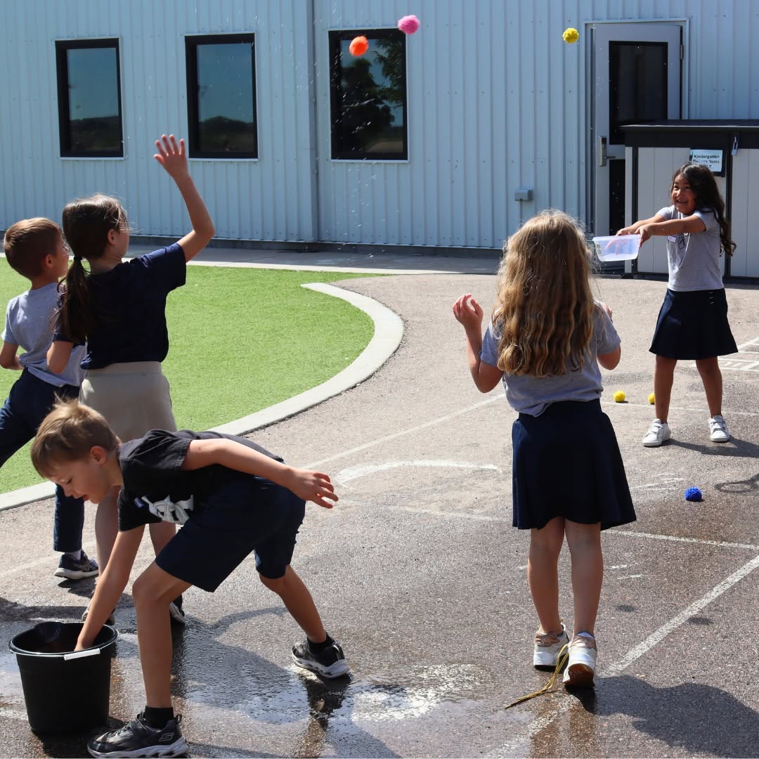 Children playing outdoors, some throwing balls, others watching, on a paved area near a building with windows.