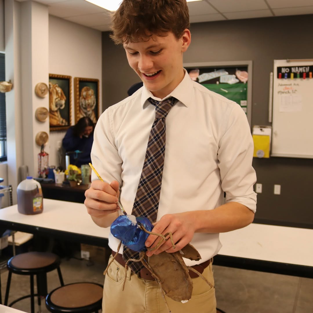 Young person smiling, holding a paintbrush and a painted object in a classroom setting.