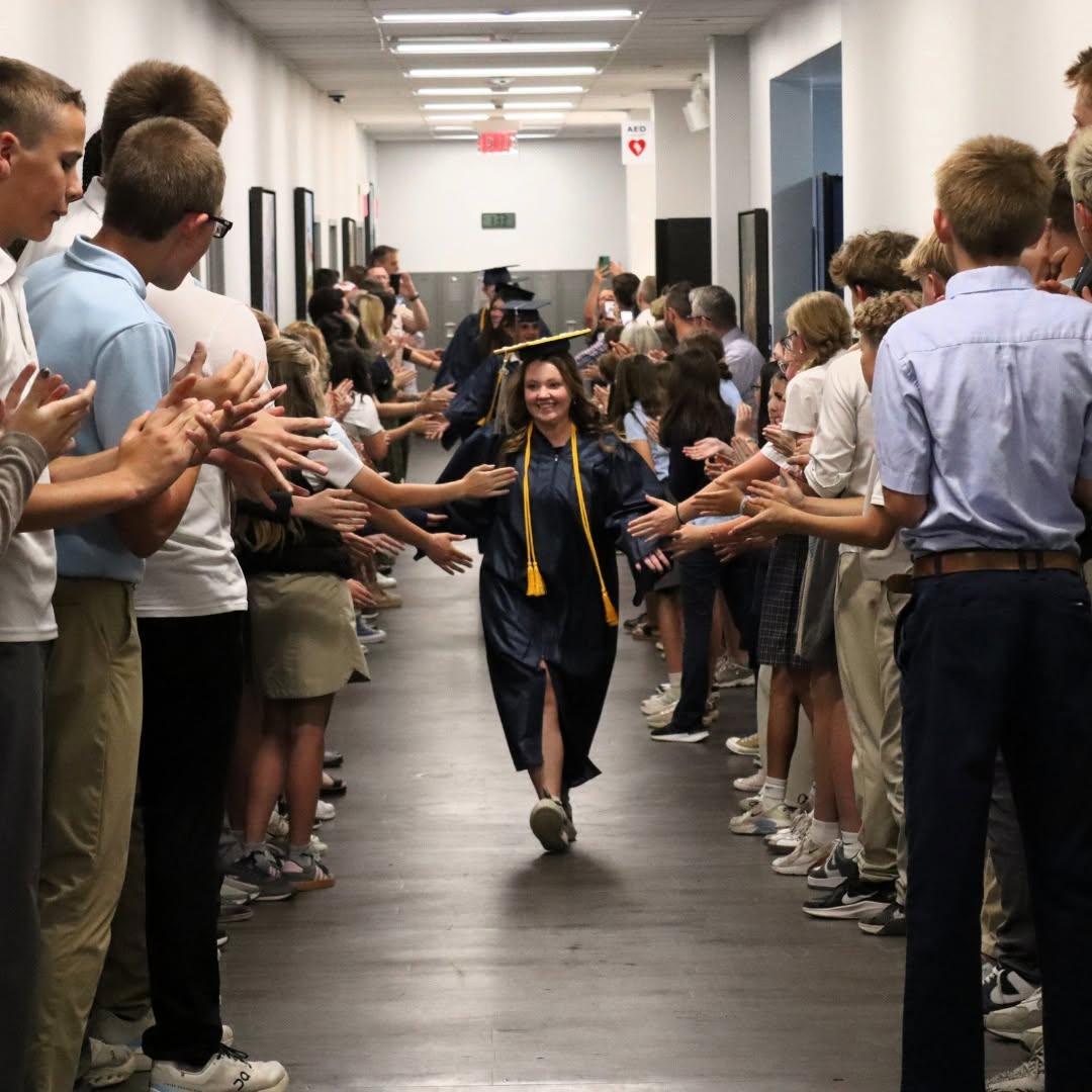 Graduate in cap and gown walking down hallway lined with applauding people on both sides.