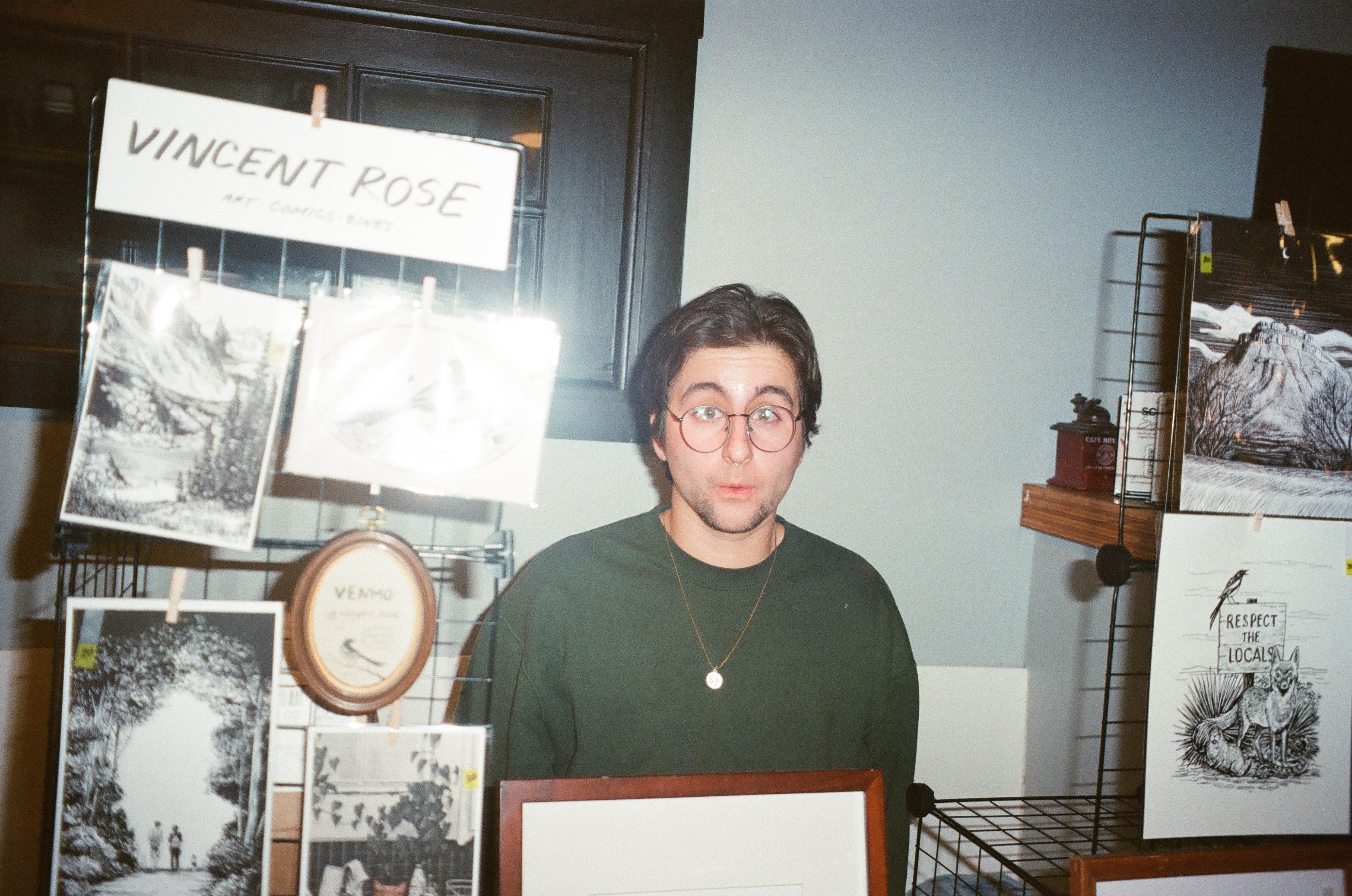 Young man with glasses and dark hair standing behind a display table at an event, with photographs and signs in the background.