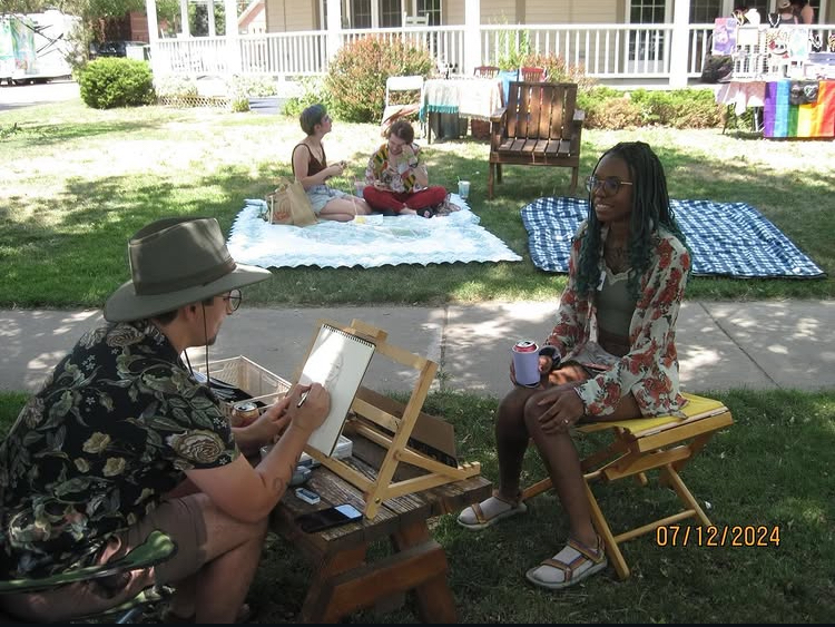 Two women sitting on benches outdoors, with children playing on a blanket in the background, under a tree.