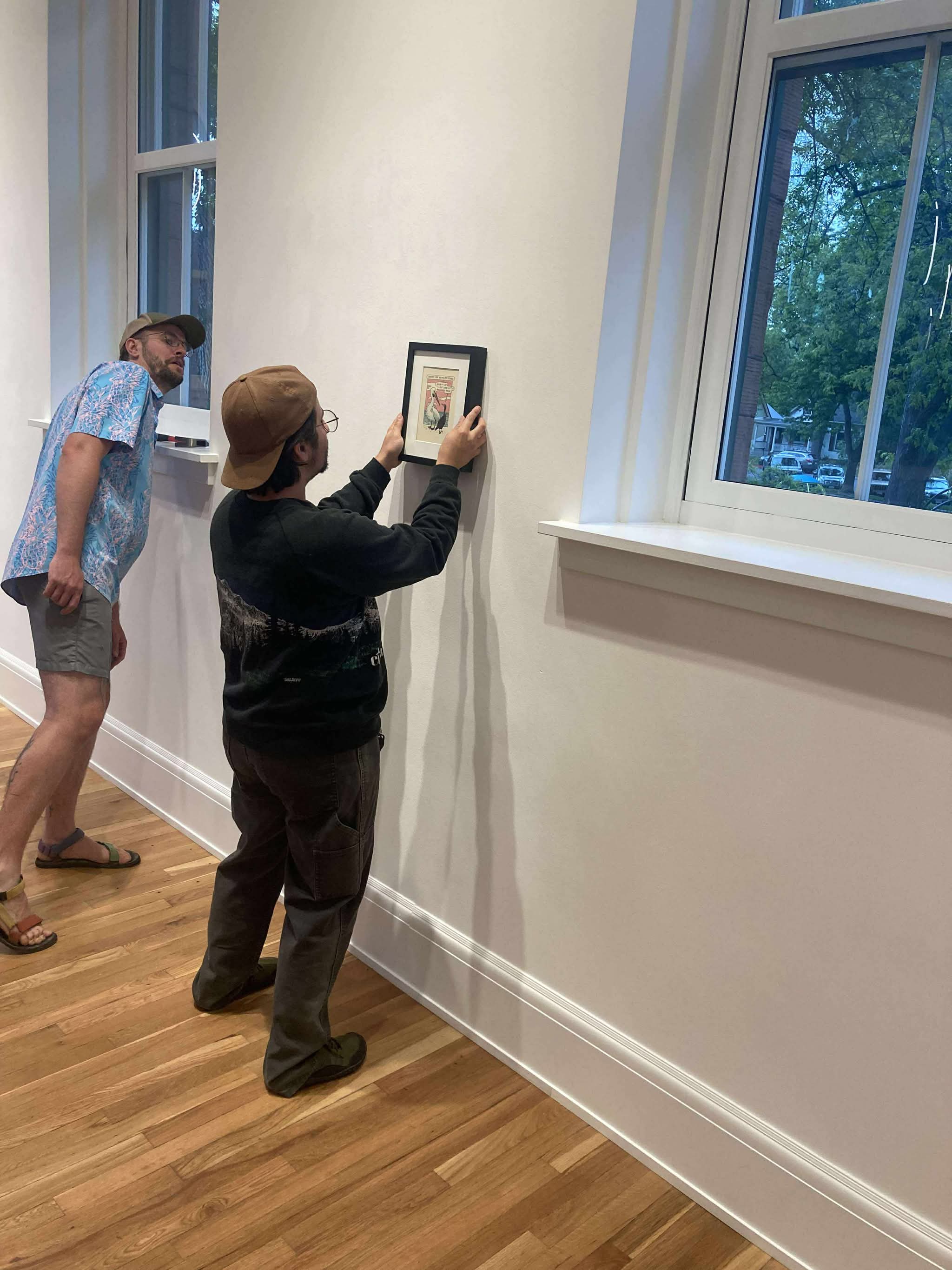 Two people in a room with wooden floor and white walls, one hanging a picture, window showing trees outside