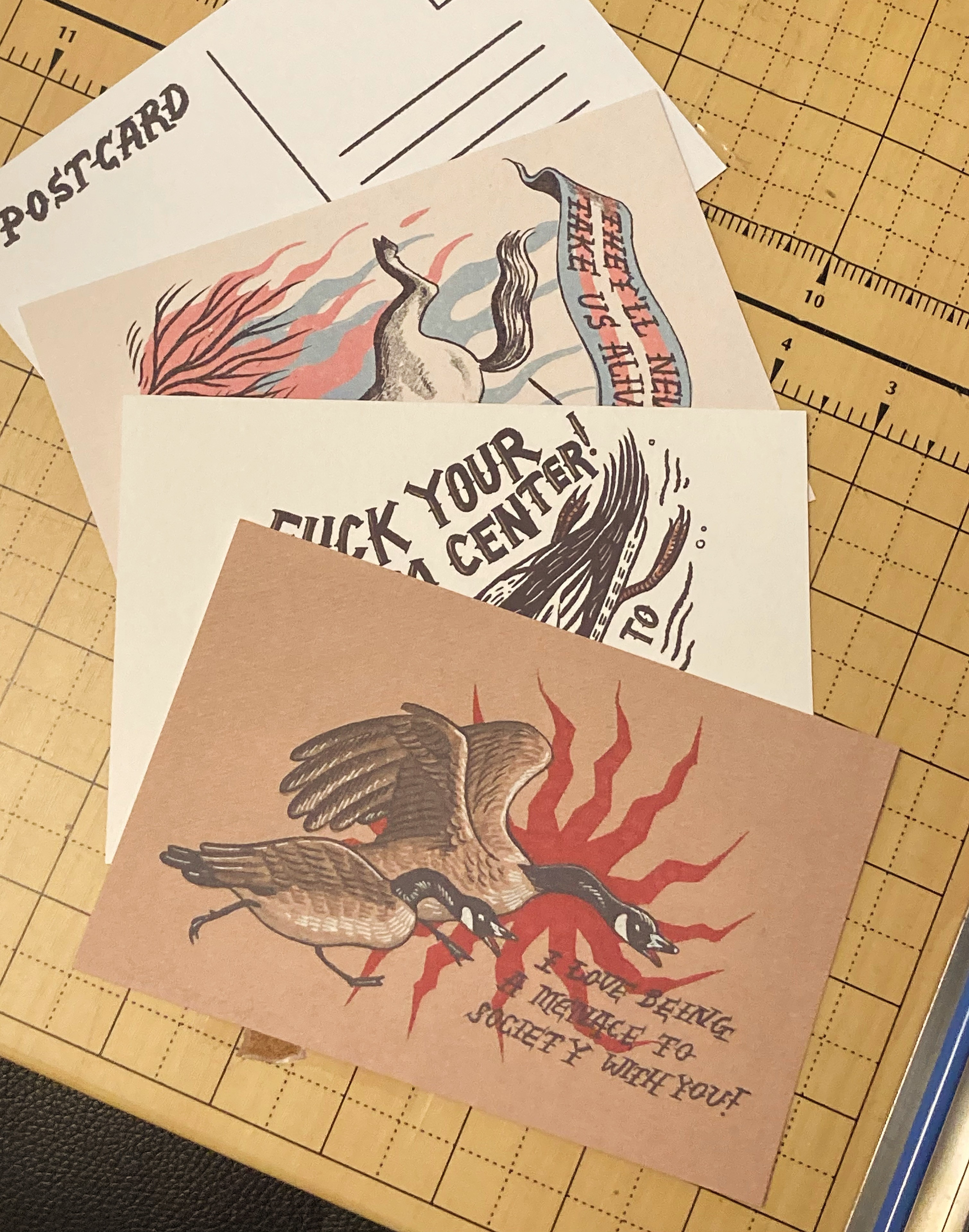 Three illustrated postcards with animals and text, placed on a yellow cutting mat with a metal ruler and pen nearby.