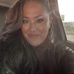 Woman with long dark hair smiling inside a vehicle, sunlight shining through window, wearing dark top.