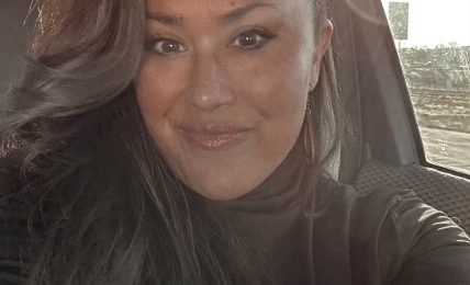 Woman with long dark hair smiling inside a vehicle, sunlight shining through window, wearing dark top.