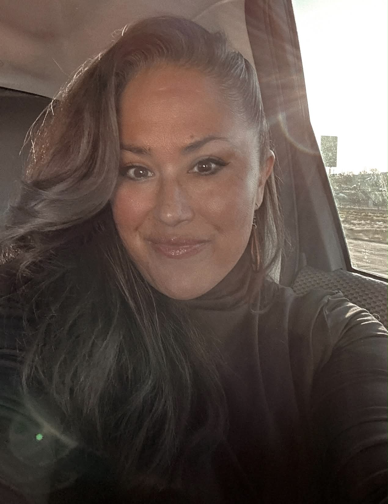 Woman with long dark hair smiling inside a vehicle, sunlight shining through window, wearing dark top.