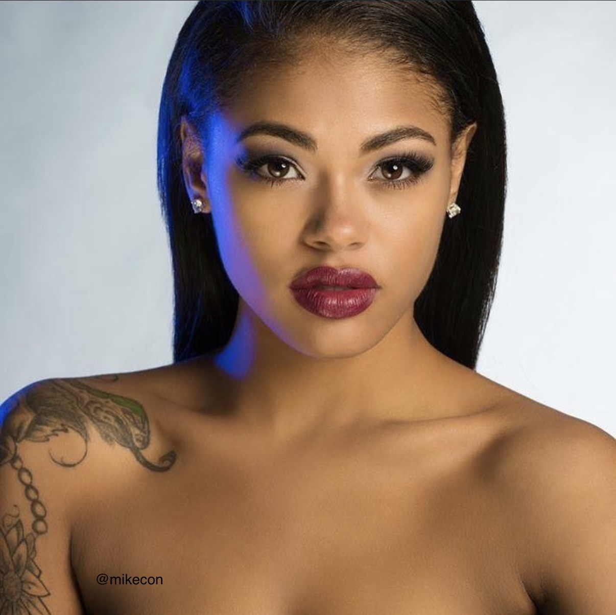 Close-up of a woman with dark hair, bold makeup, and a tattoo on her shoulder, looking directly at the camera.