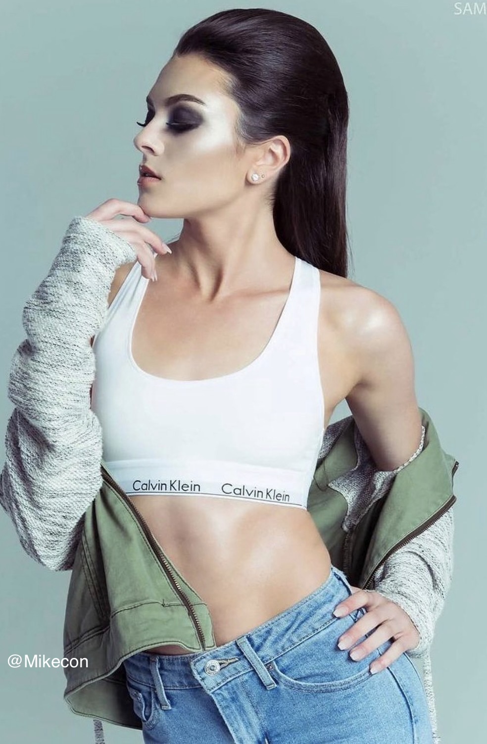 Woman with dark hair, makeup, wearing a white Calvin Klein sports bra, green jacket, and jeans, posing sideways.