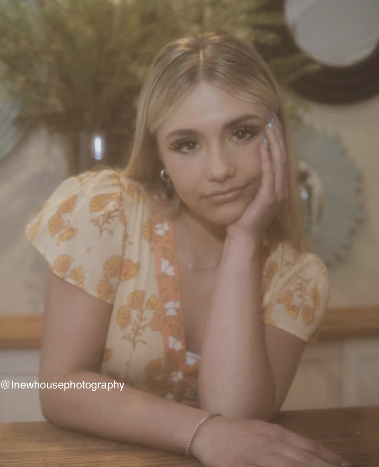Young woman with blonde hair resting her face on her hand, wearing a floral dress, sitting at a table.