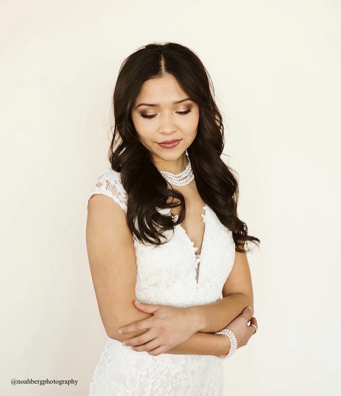 Young woman with long dark hair in a white lace dress, arms crossed, looking down, neutral background.