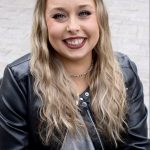 Young woman with long wavy blonde hair smiling, wearing a black leather jacket and gray top, outdoors.