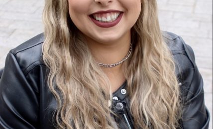 Young woman with long wavy blonde hair smiling, wearing a black leather jacket and gray top, outdoors.