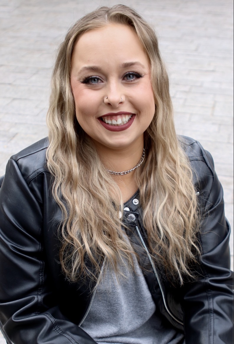 Young woman with long wavy blonde hair smiling, wearing a black leather jacket and gray top, outdoors.