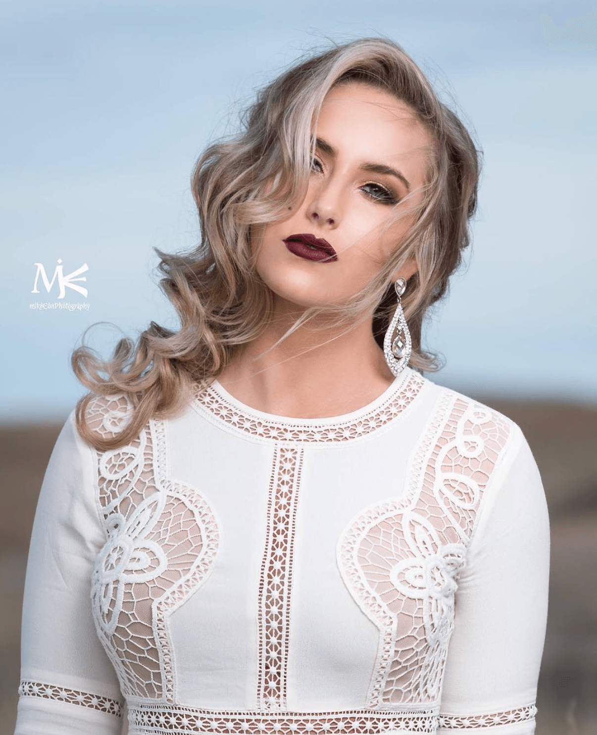 Woman with wavy blonde hair and dark lipstick wearing a white lace dress and large earrings outdoors.
