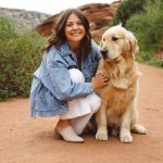 Woman crouching next to a golden retriever on a dirt path with greenery and rocks in the background.