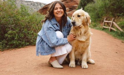 Woman crouching next to a golden retriever on a dirt path with greenery and rocks in the background.
