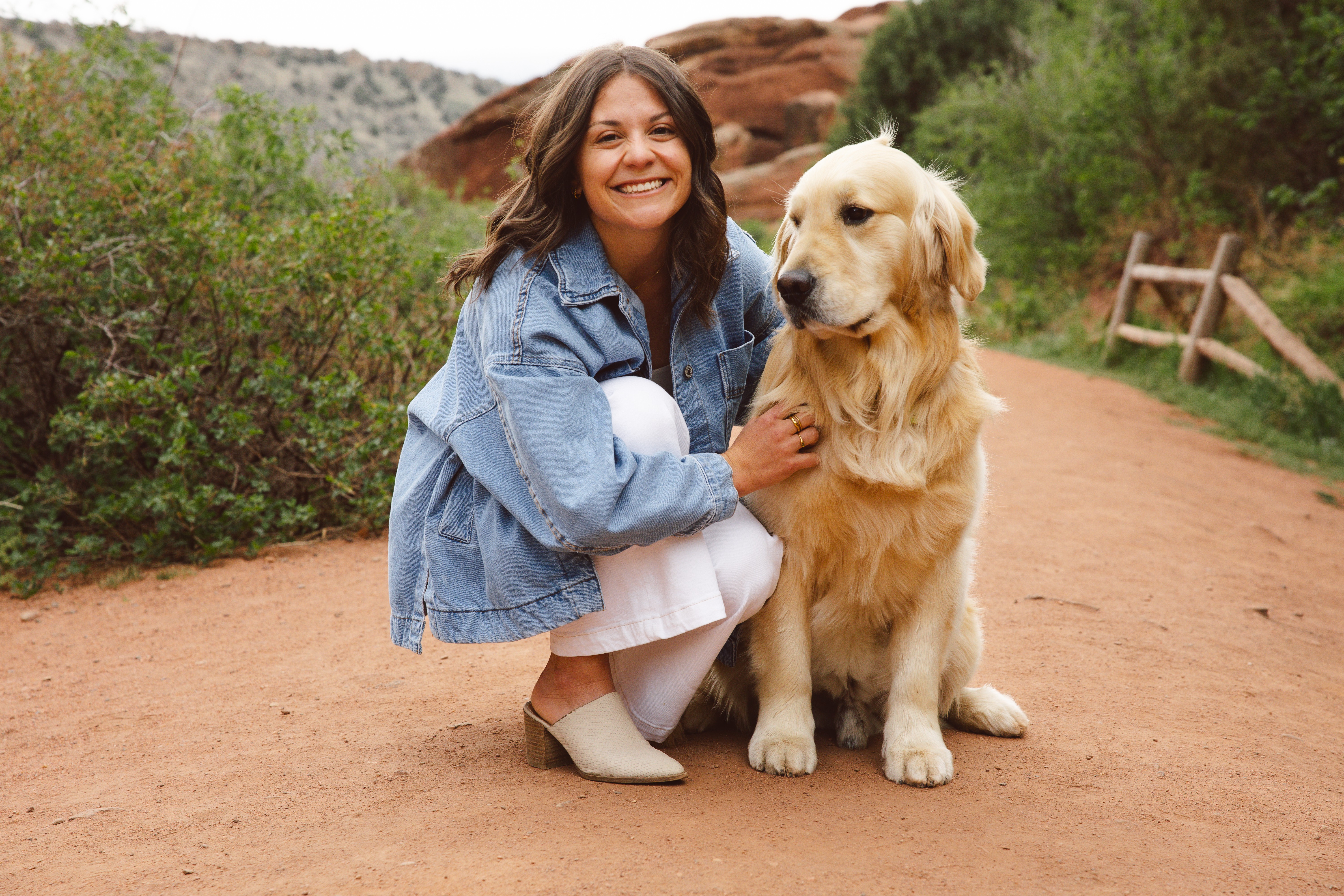 Woman crouching next to a golden retriever on a dirt path with greenery and rocks in the background.