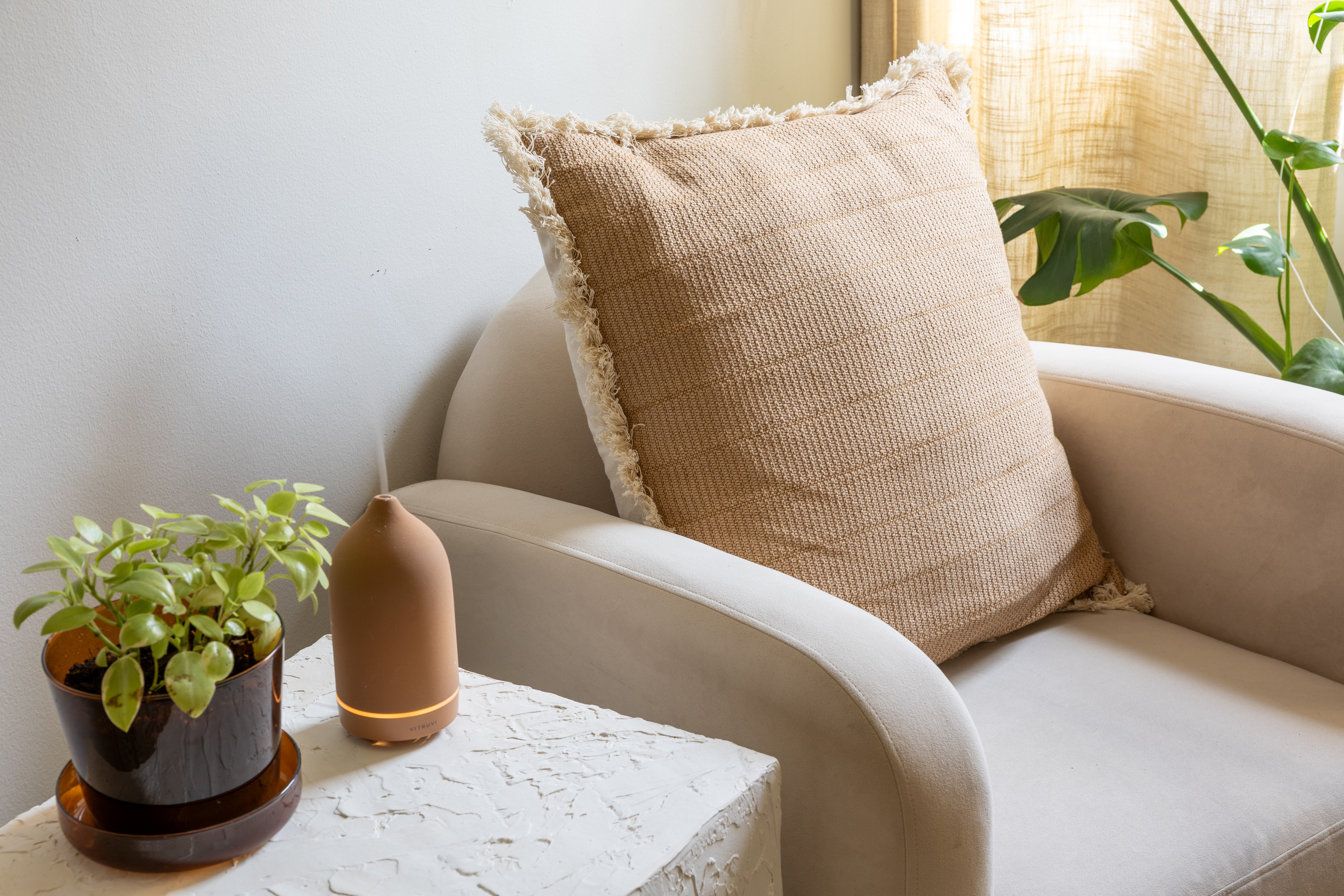 A beige cushion on a white armchair next to a window with a plant, a small potted plant, and a decorative object on a table.