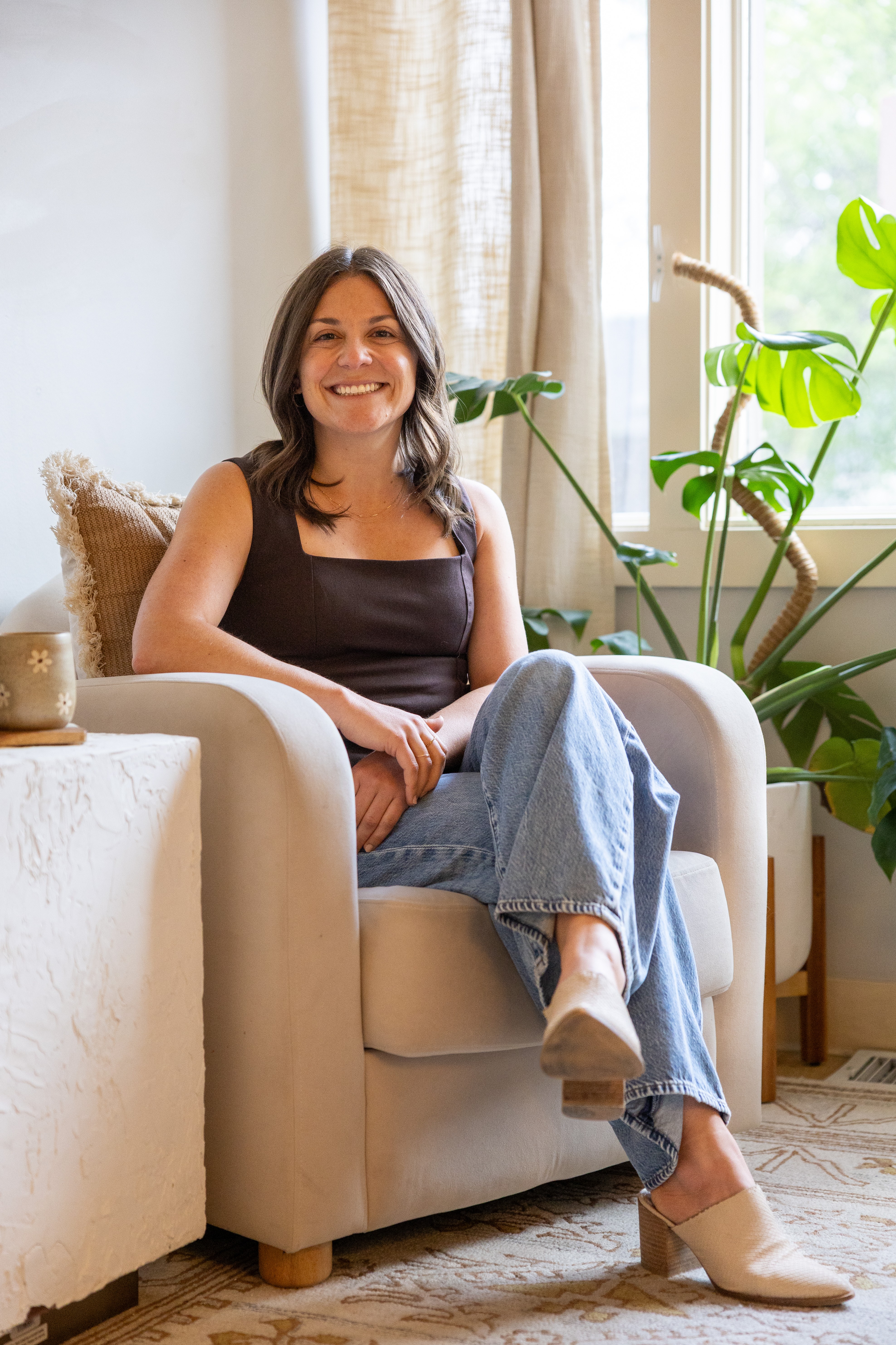 Woman sitting on a beige armchair, smiling, in a bright room with large windows and green plants.