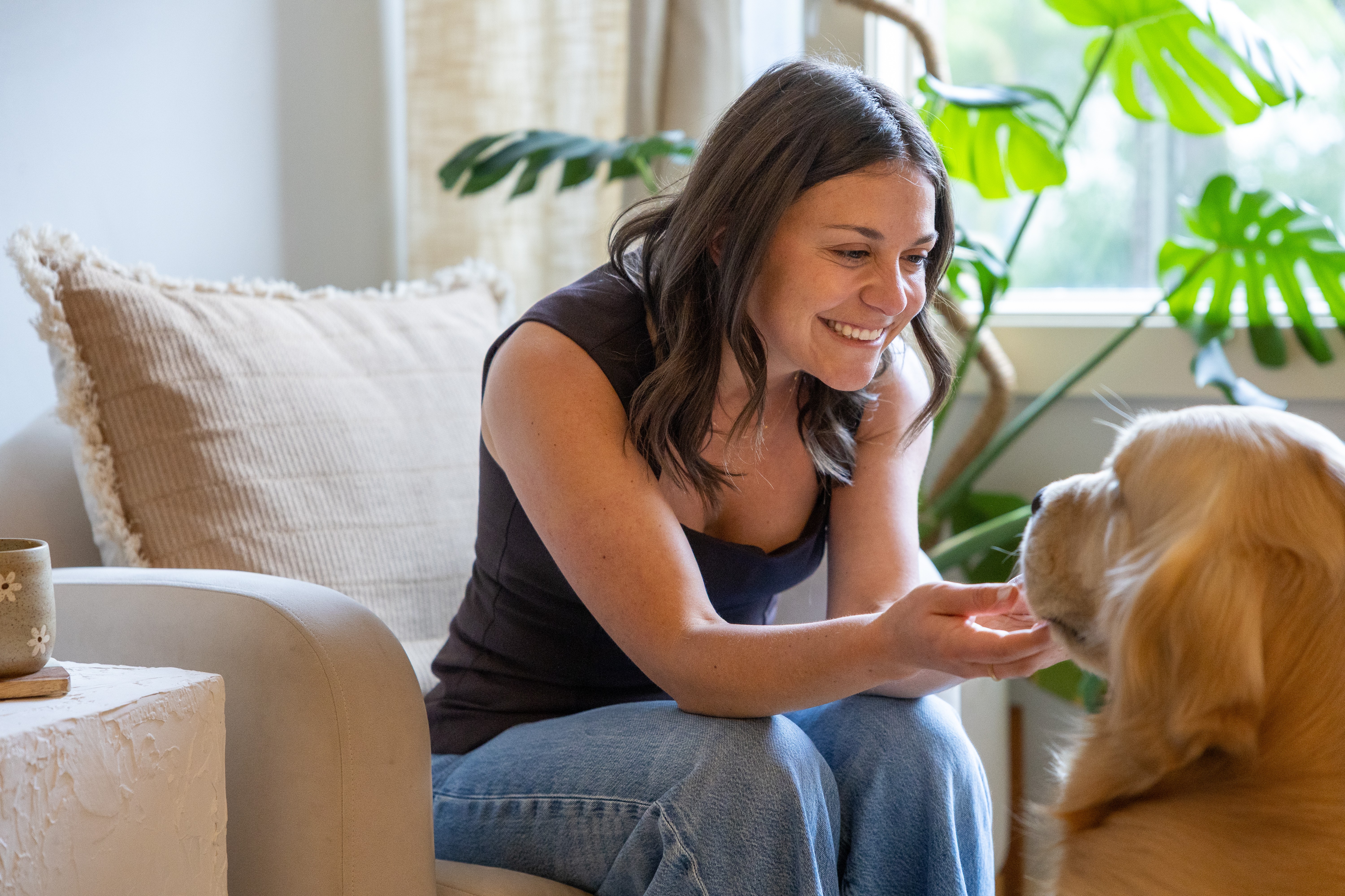 Woman smiling and petting a dog indoors with plants and a sofa in the background.