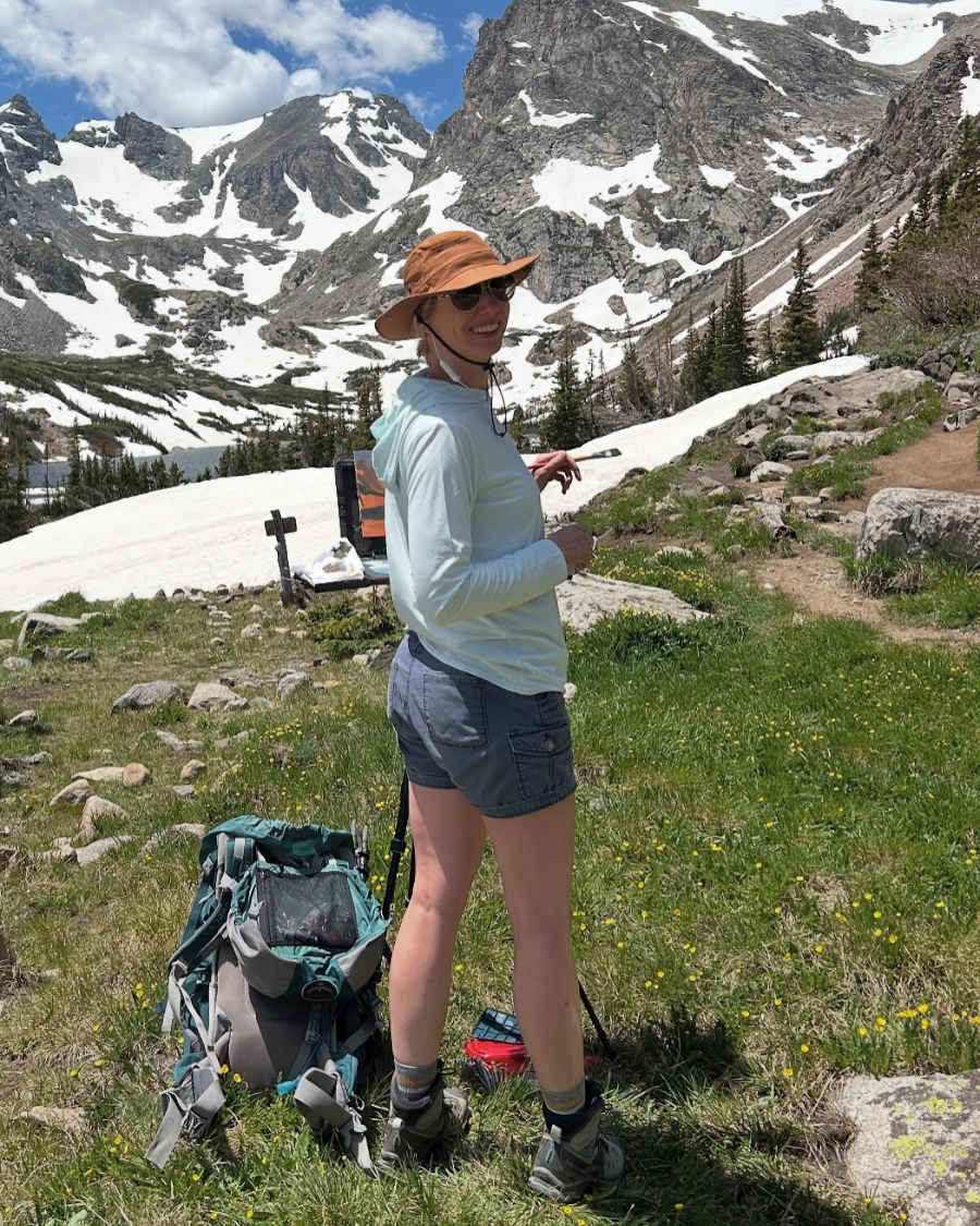 Person standing outdoors in a mountainous area with snow, wearing a hat, sunglasses, light jacket, and shorts, with a backpack nearby.
