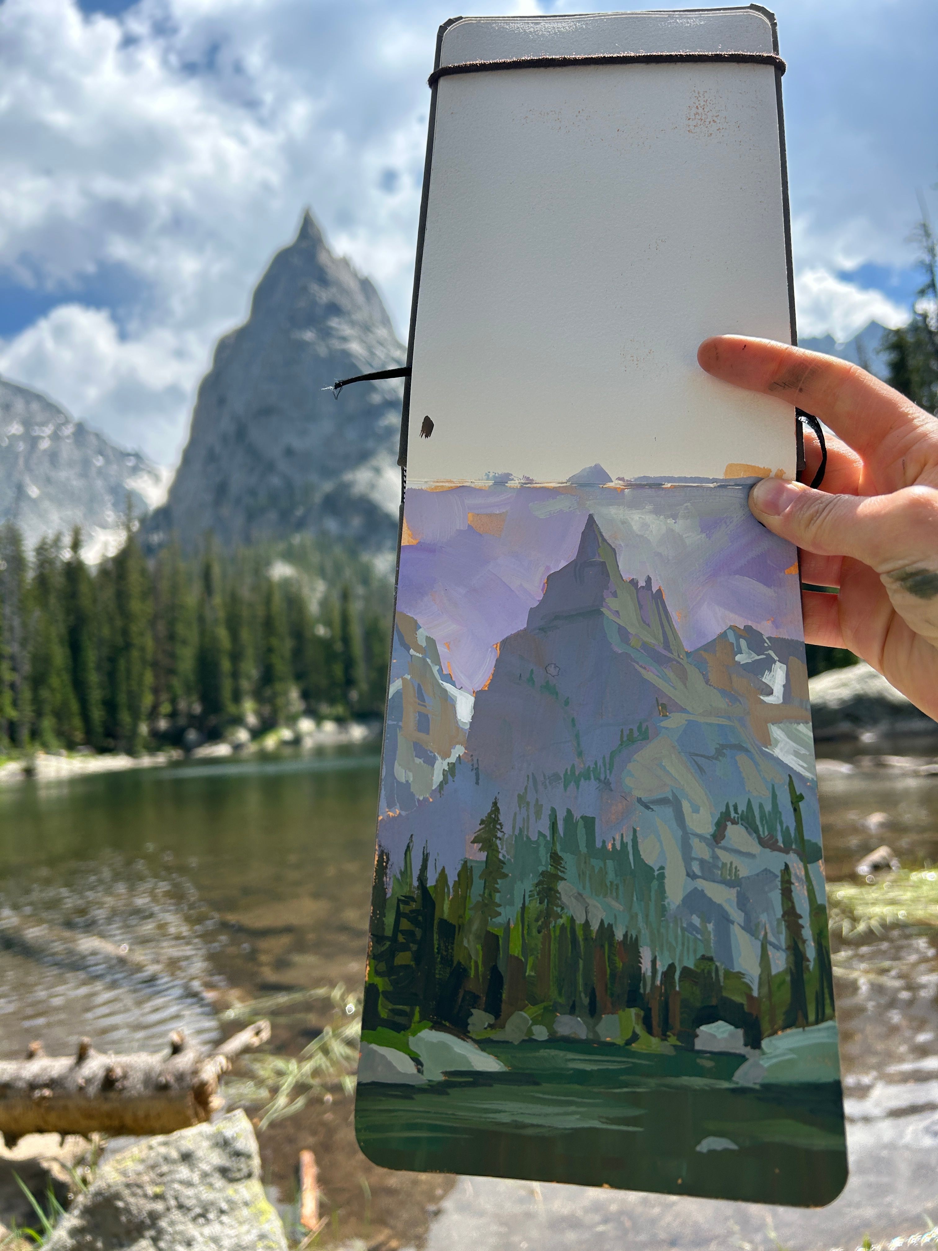 Person holding a painting of mountains and trees near a river with mountains in the background.