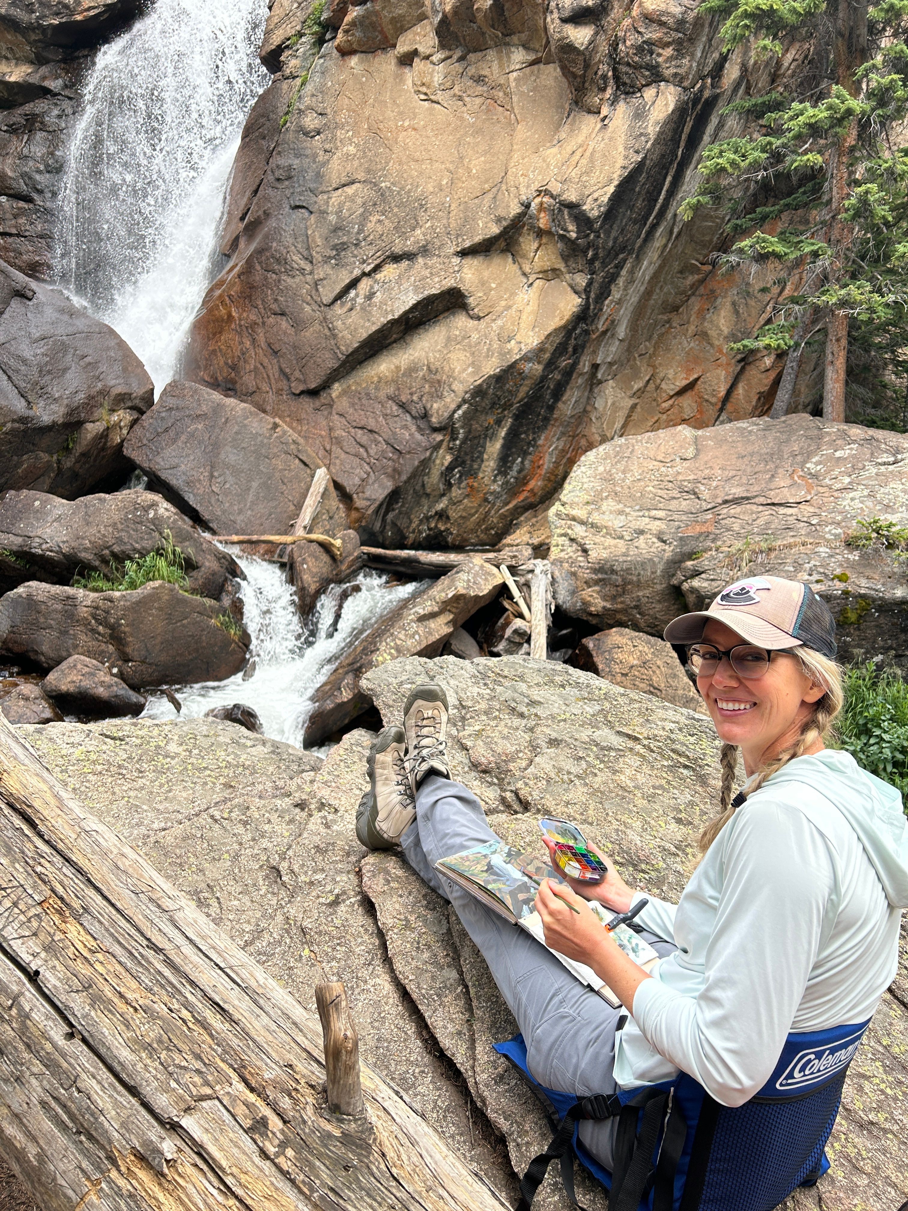 Person crouching on rocks near a waterfall, wearing a hat, sunglasses, and light-colored clothing, holding a camera or phone.