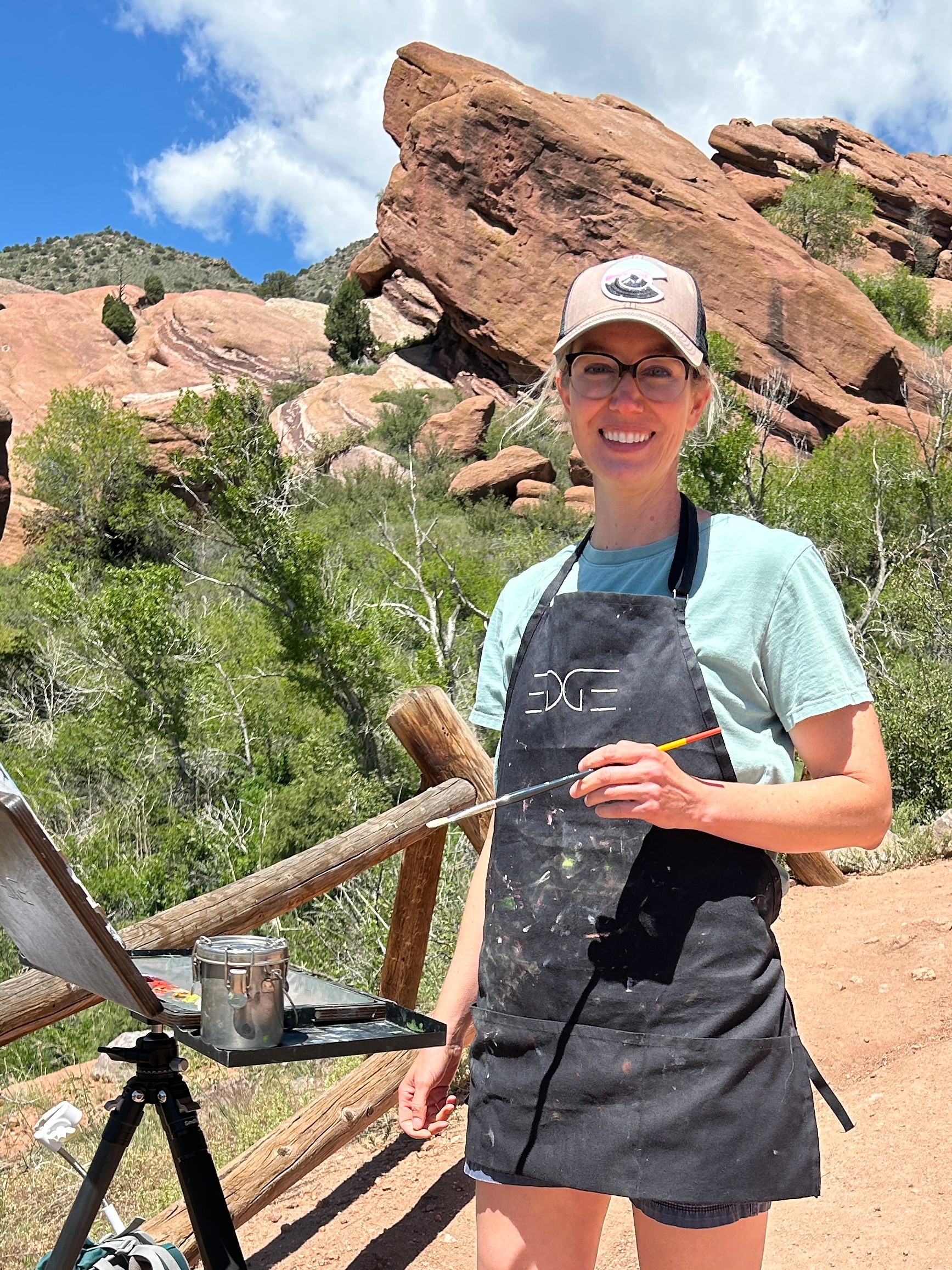 Young woman outdoors with large rock formations and greenery, wearing a cap, apron, and glasses, smiling at camera.