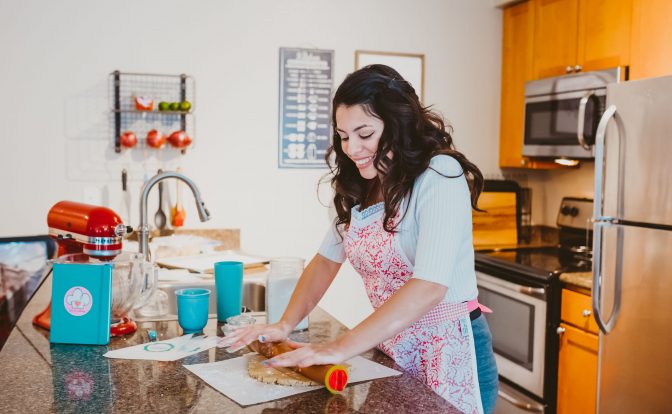Woman in a kitchen preparing food at a counter with various kitchen items around her.