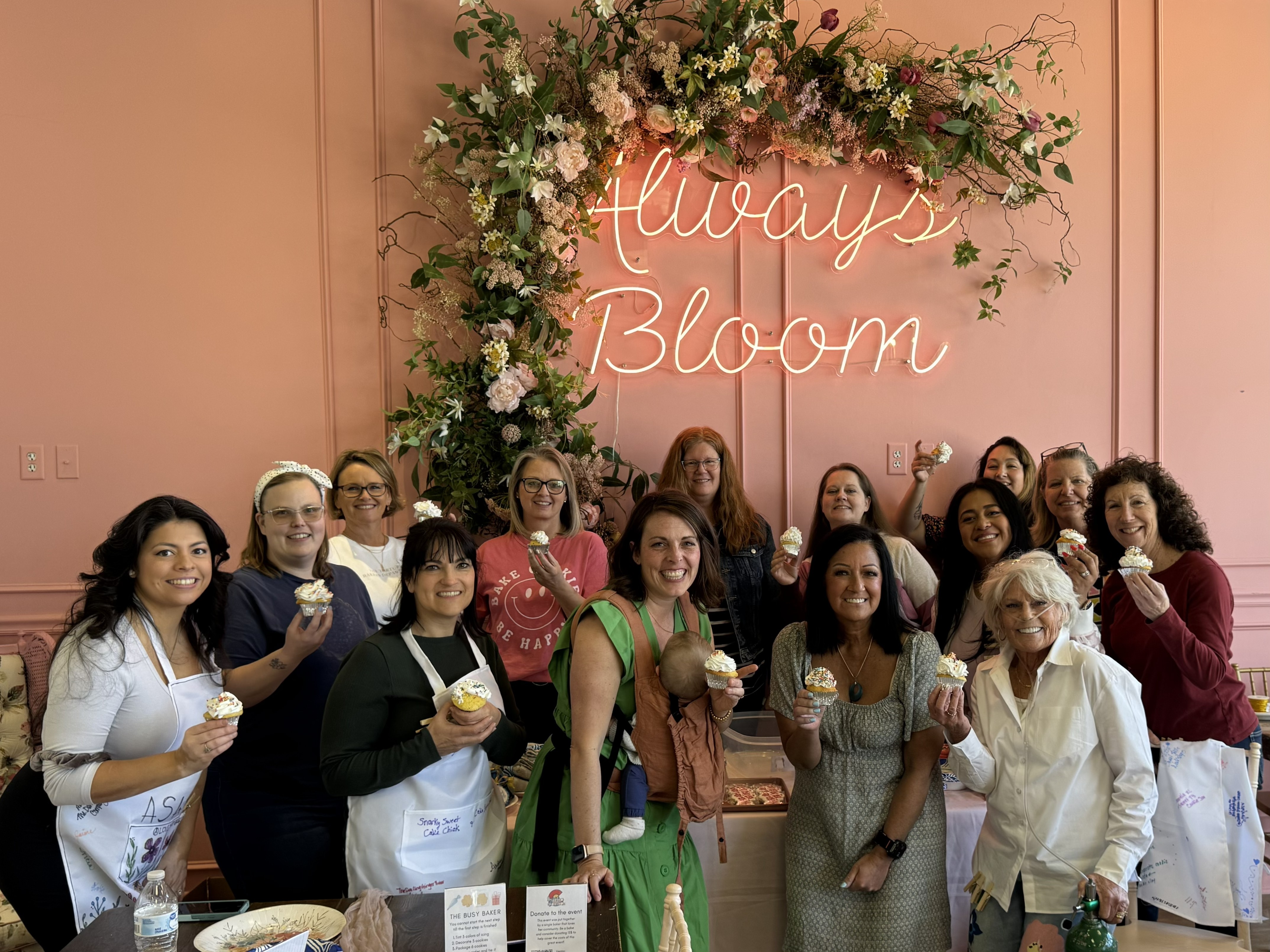 Group of women standing together in front of a floral display with a neon sign that says 'Always Bloom', holding food or drinks.