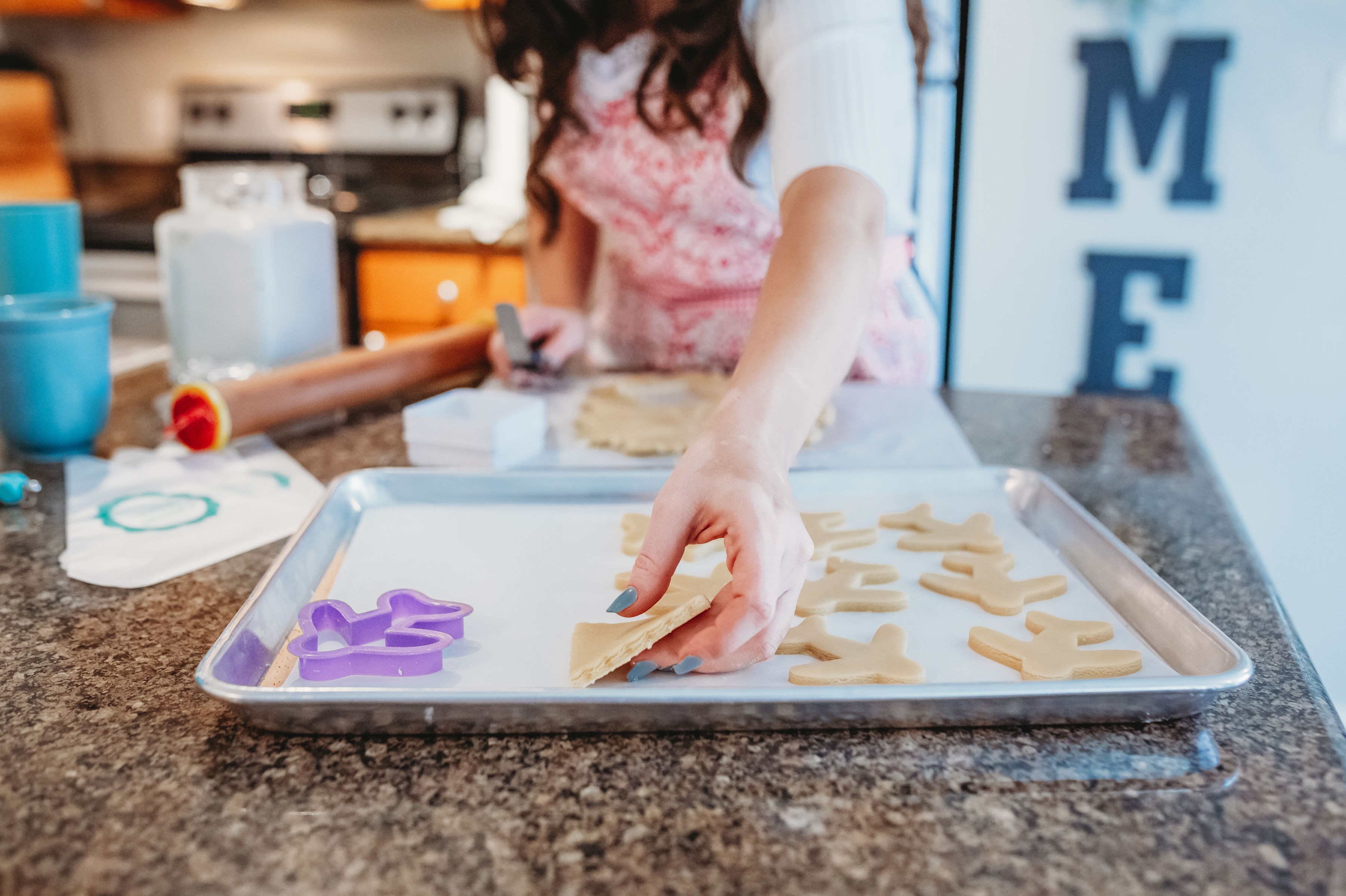 Person decorating cookies on a baking sheet with cookie cutters, with kitchen items in background.