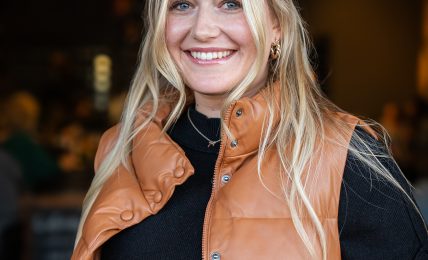 Smiling woman with long blonde hair wearing a black top and tan vest, indoors with blurred background.