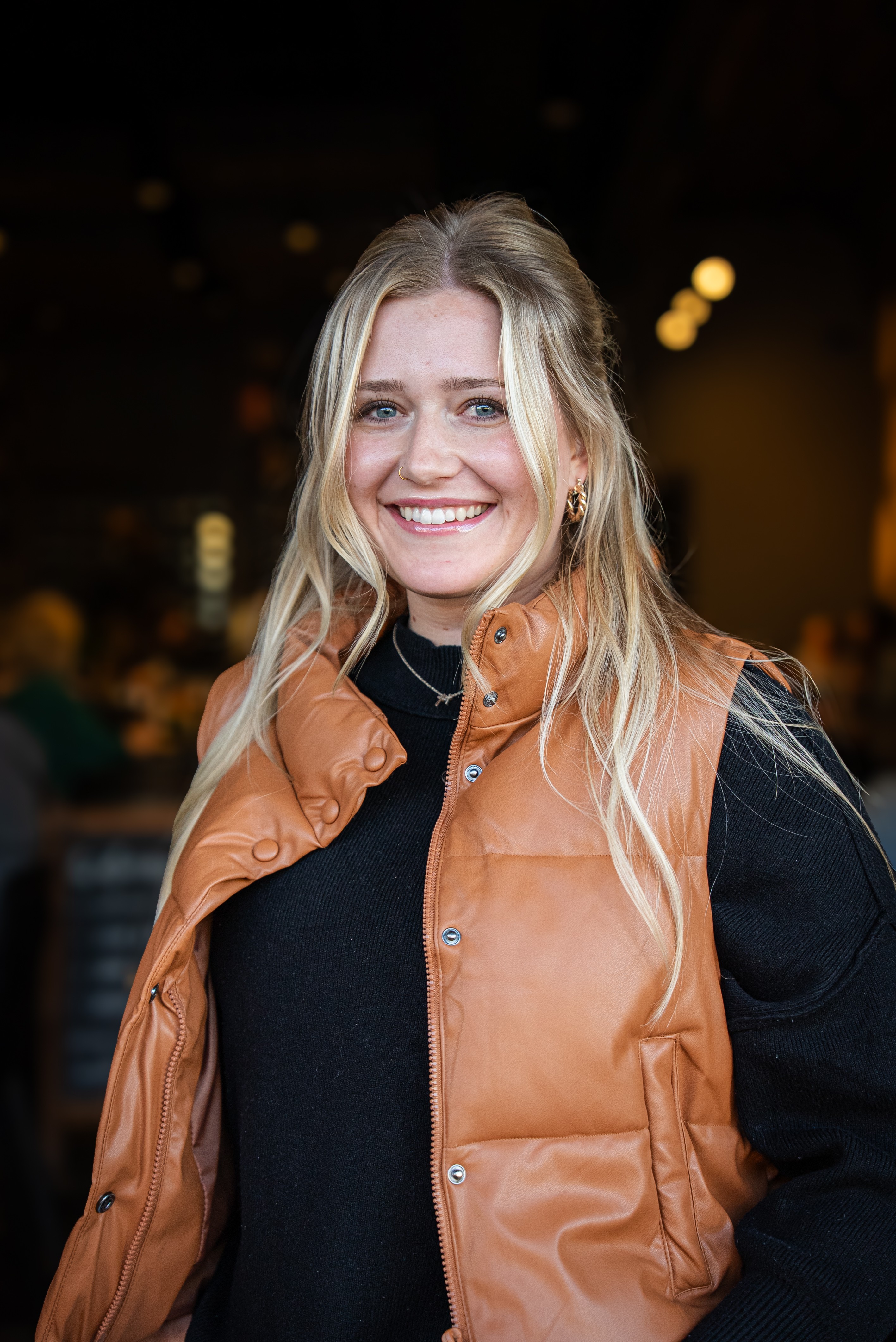 Smiling woman with long blonde hair wearing a black top and tan vest, indoors with blurred background.
