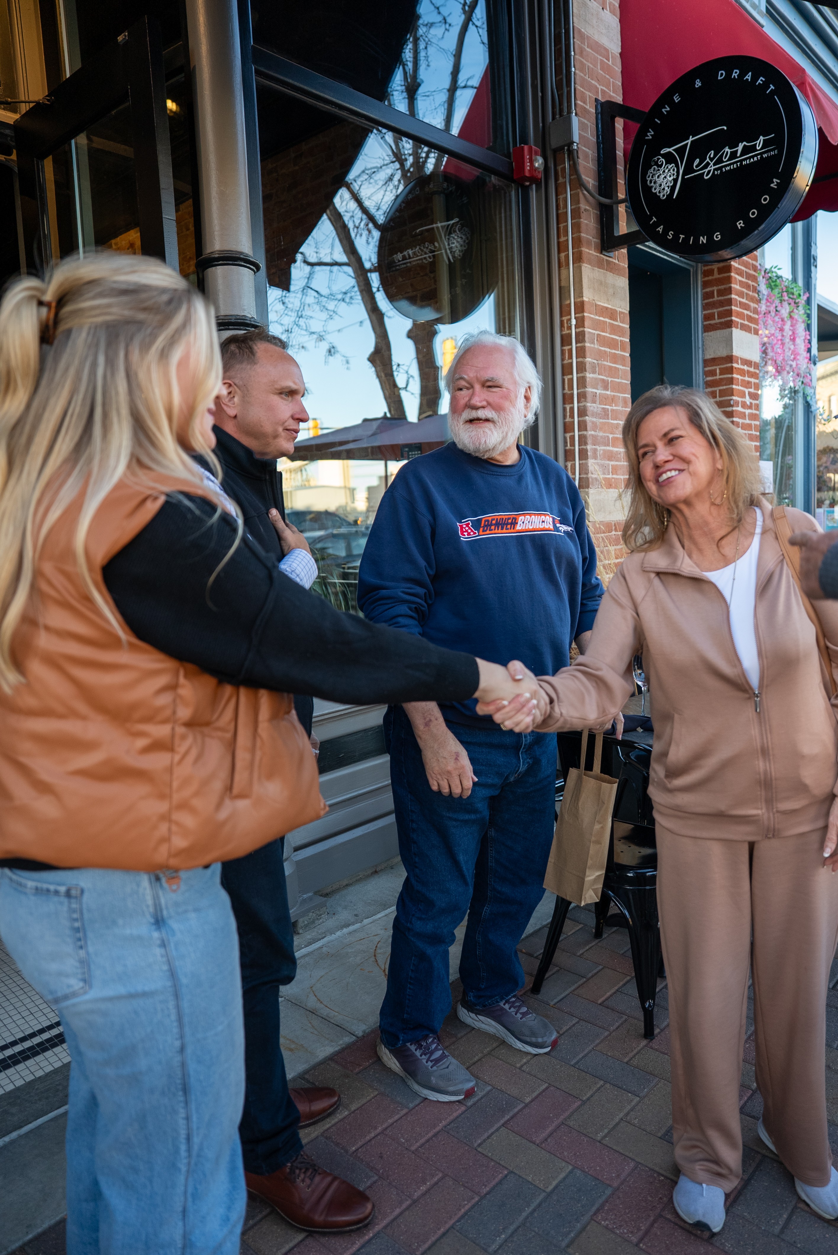 Four people shake hands outside a building with a sign, smiling and engaging in conversation.