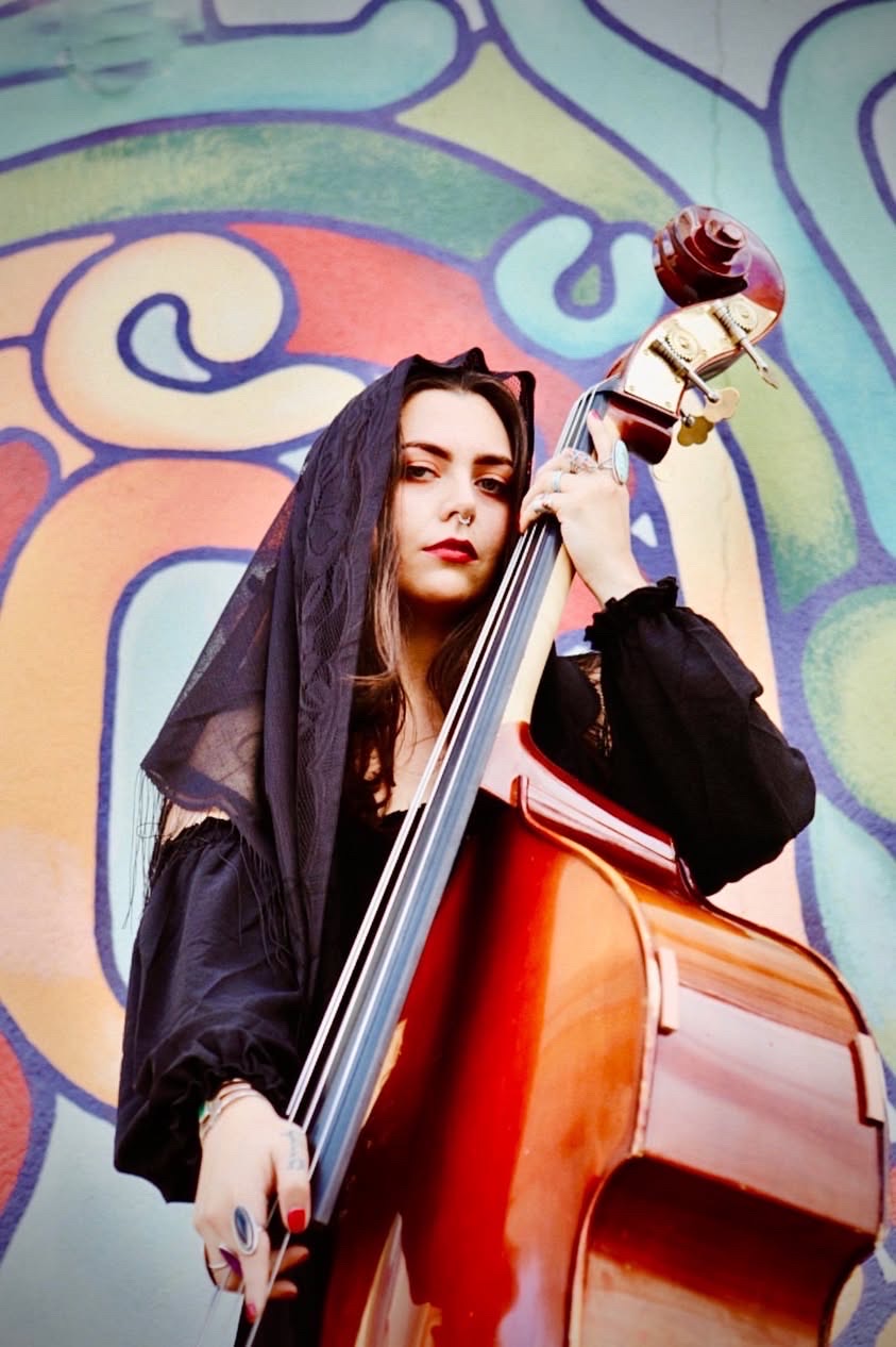 Young woman with long dark hair holding a double bass against a colorful abstract background.